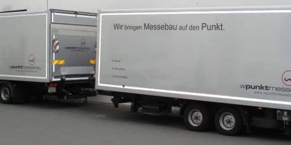 Zwei Lastwagen mit eleganter silberner Außenfassade stehen nebeneinander. Auf einem steht: „Wir bringen Messebau auf den Punkt.“ Die Transportfahrzeuge gehören der Firma „wpunktmessebau“, die sich auf präzisen Messebau spezialisiert hat.