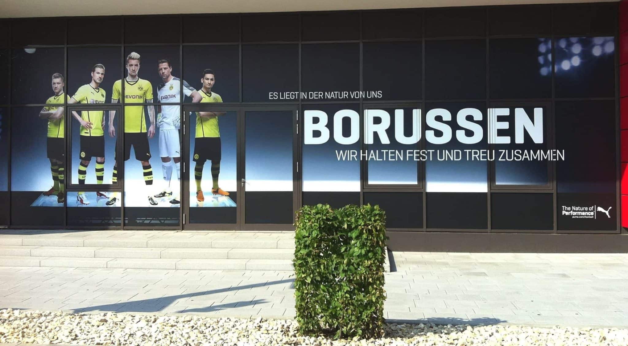 Ein Schaufenster zeigt eine große Werbung mit Fußballspielern in gelb-schwarzen Trikots, begleitet von deutschem Text. Ein kleiner grüner Busch verleiht der Vorderseite Charme, während der Innenausbau mit einem Kieselsteinbereich die einladende Atmosphäre des Ladens verstärkt.