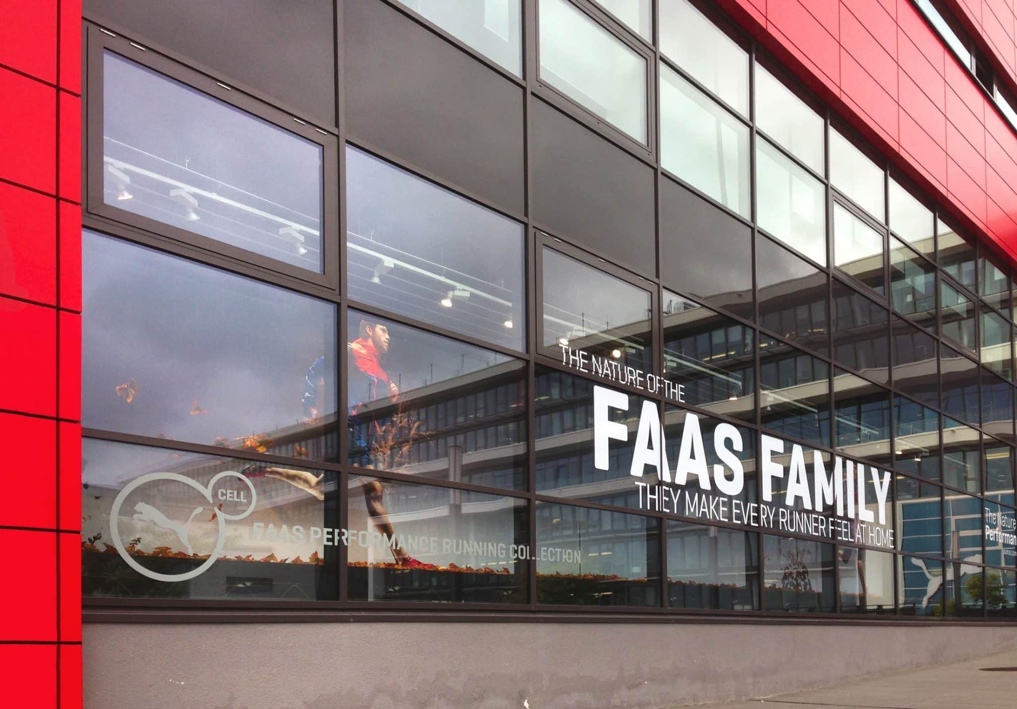 Ein modernes Gebäude mit großen Fenstern präsentiert Werbung für die Laufkollektion „Faas Family“. Im Inneren ist Sportbekleidung zu sehen, was den Fokus auf den Innenausbau unterstreicht. Die Außenfassade ist überwiegend rot gehalten, auf dem Glas spiegeln sich Himmel und umliegende Gebäude.