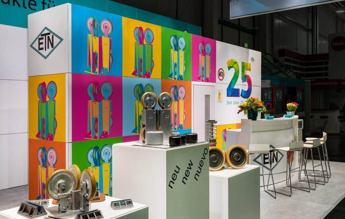 Messebau Augsburg – Interlift Messestand von ETN mit bunten Würfeln, Industrie-Rollen und moderner Theke.