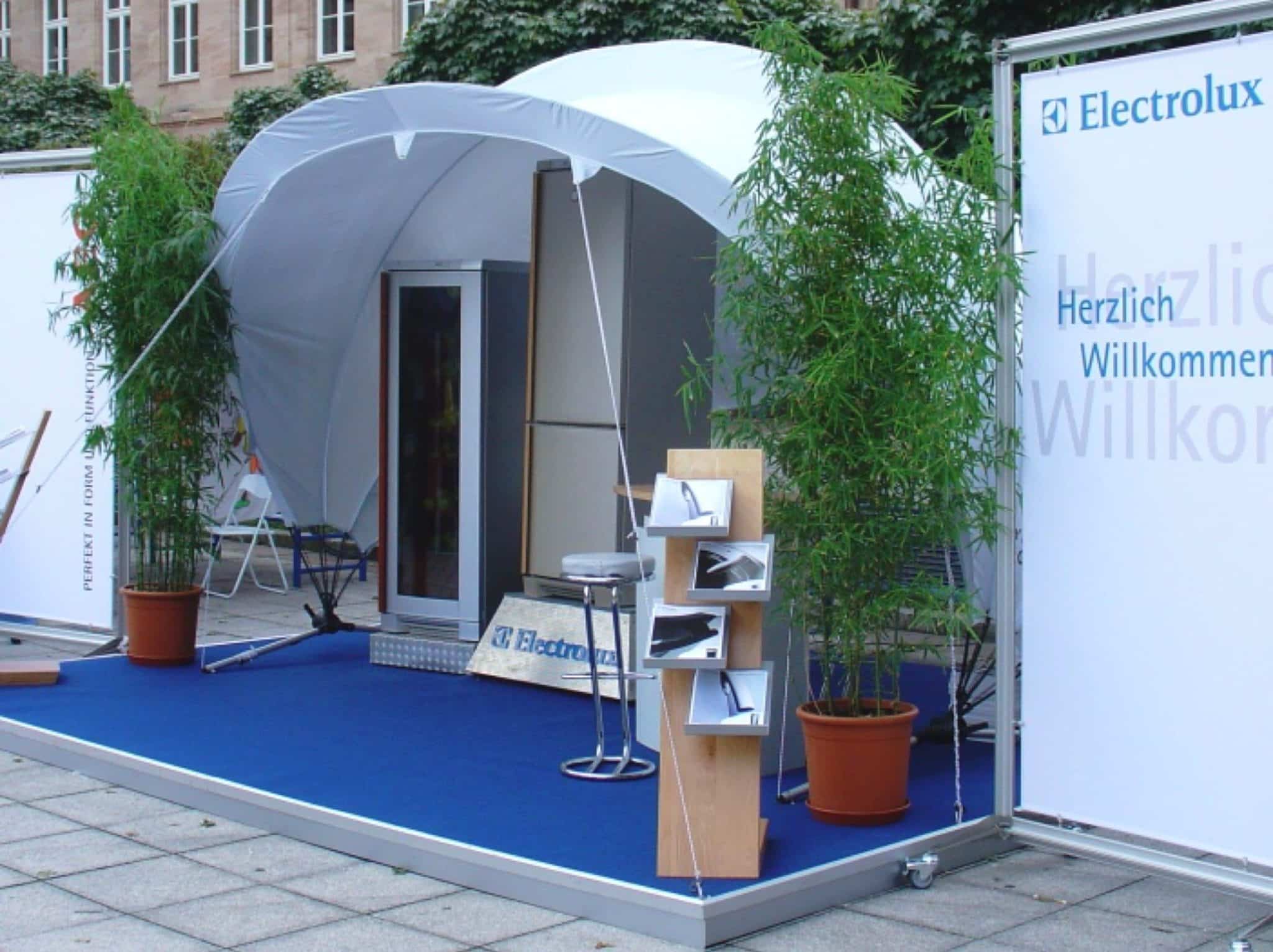 Ein kleiner Messestand mit weißem Vordach, blauem Teppich und Electrolux-Branding steht für Veranstaltungen bereit. Zwei hohe Topfpflanzen flankieren den Eingang und laden die Gäste ein. Vor dem Stand befindet sich ein Stand mit Broschüren, der leicht zugänglich ist.