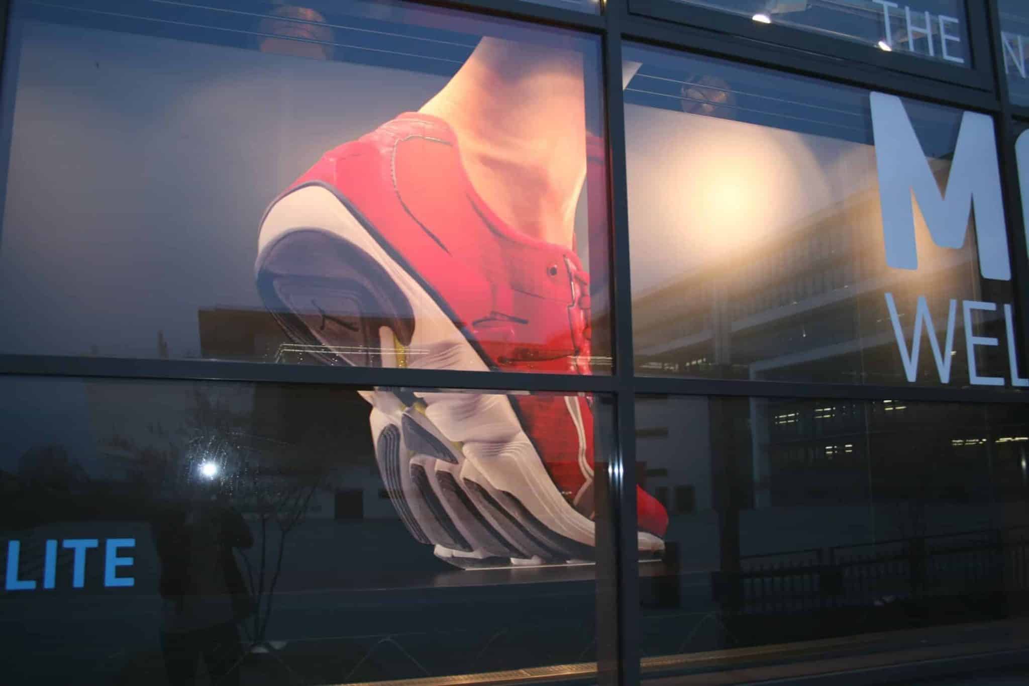 Schaufensterdisplay mit dem großen Bild eines roten Sportschuhs mit dynamischer, übertriebener Sohle. Der Schuh scheint in Bewegung und ergänzt perfekt die elegante Innenkonstruktion, während Reflexionen und Gebäudeelemente über das Glas tanzen.