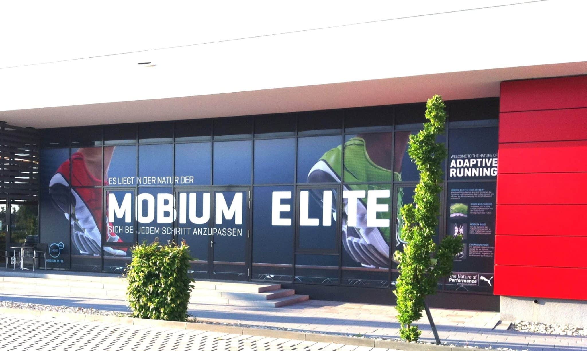 Schaufenster mit großen Fenstern, in denen in fetten Buchstaben der Schriftzug „Mobium Elite“ zu sehen ist. Die Inneneinrichtung betont Bilder von Laufschuhen und einen Slogan zum adaptiven Laufen, während im Vordergrund ein kleiner Baum thront.