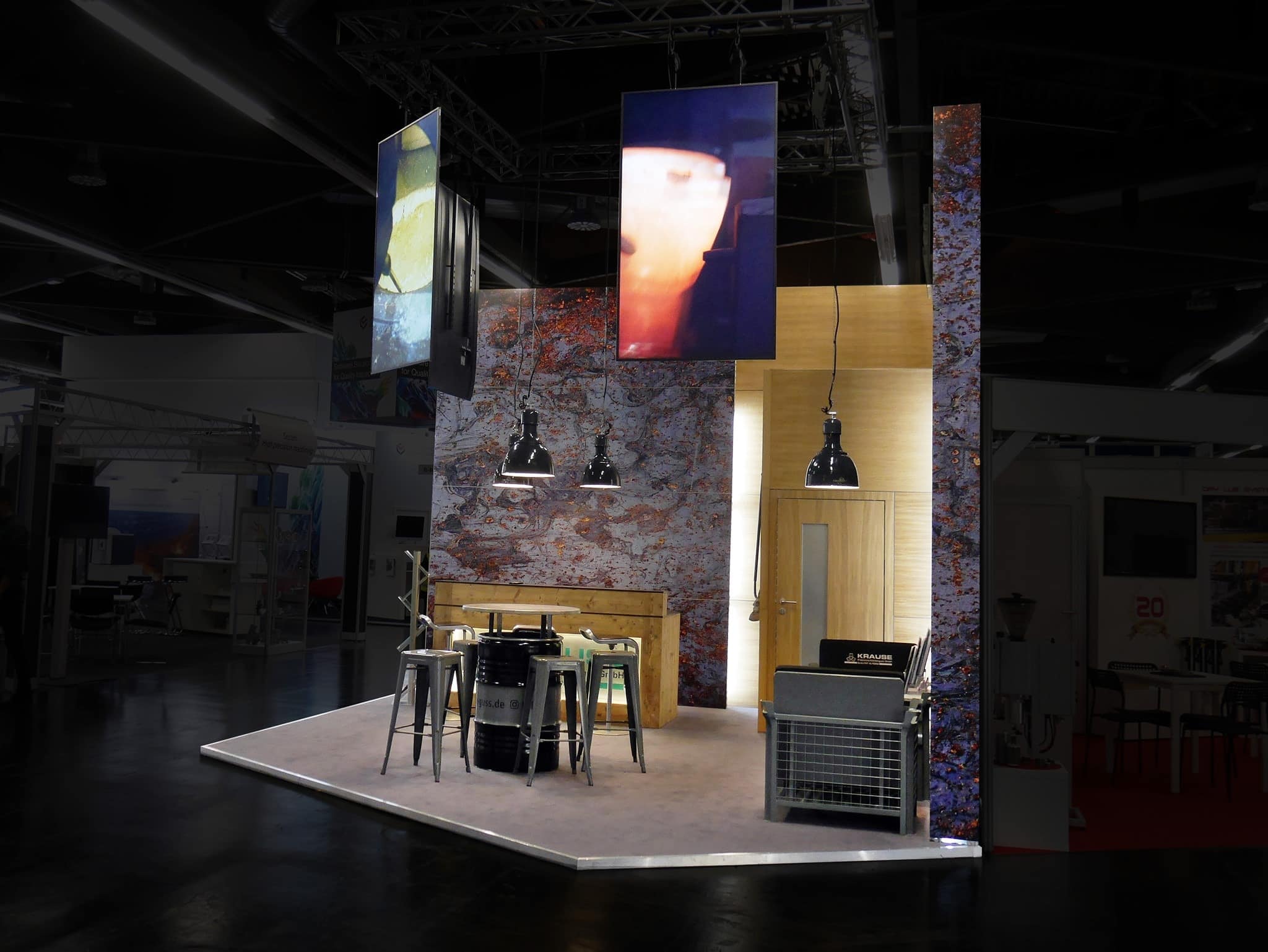 Ein moderner Messestand mit einer kleinen, eleganten Kücheneinrichtung. Er verfügt über eine Theke mit Metallstühlen, hängenden Industrieleuchten, abstrakter Wandkunst und Küchengeräten. Im Hintergrund befindet sich eine schwach beleuchtete Messehalle.