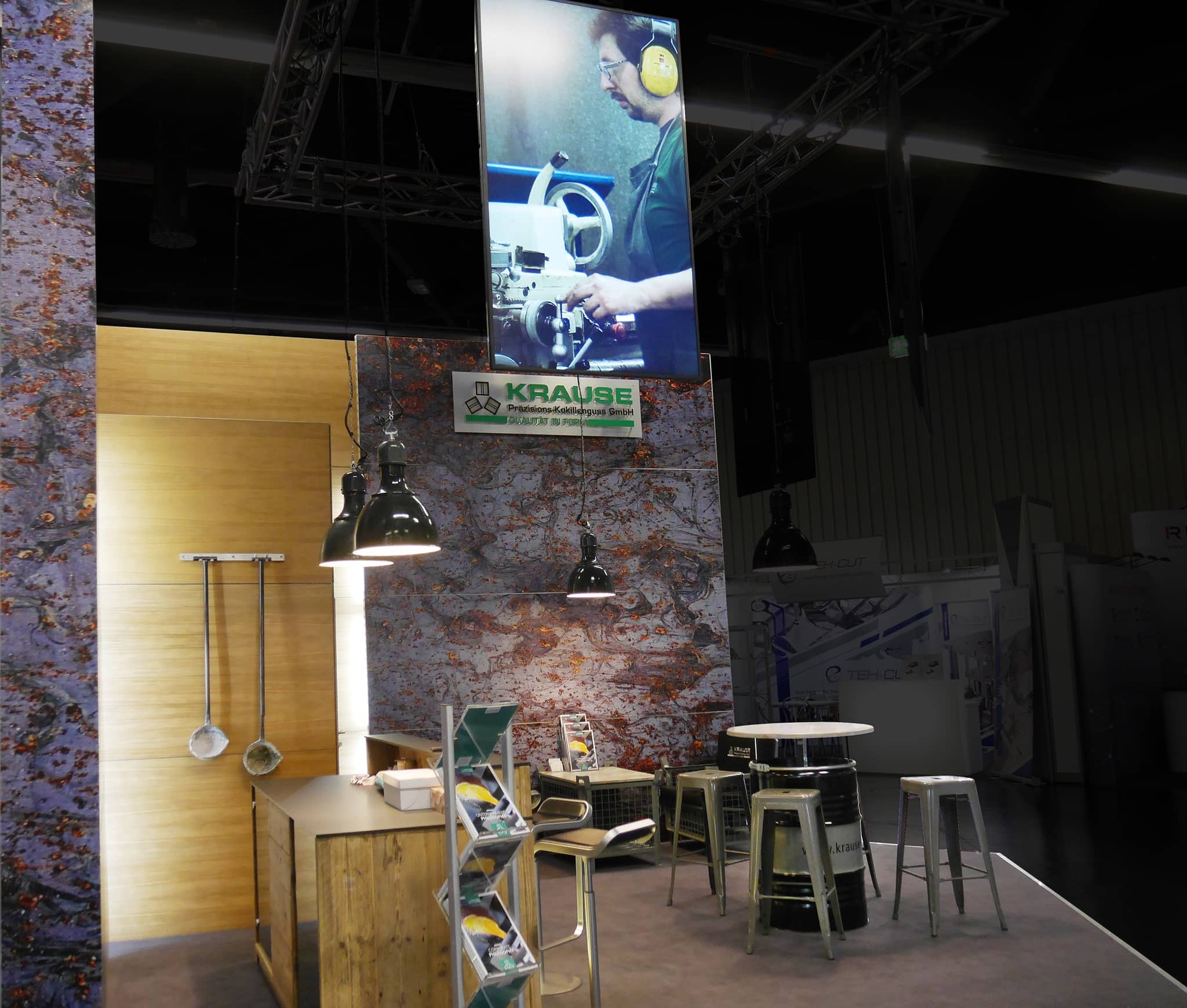Ein Messestand mit einem Schild, das eine Person an einer Maschine zeigt, einem Krause-Display, einer Holztheke mit Hockern und Beleuchtung im Industriestil. Broschüren werden auf einem Ständer ausgestellt, und der Stand ist im rustikalen Metall- und Holzdesign gestaltet.