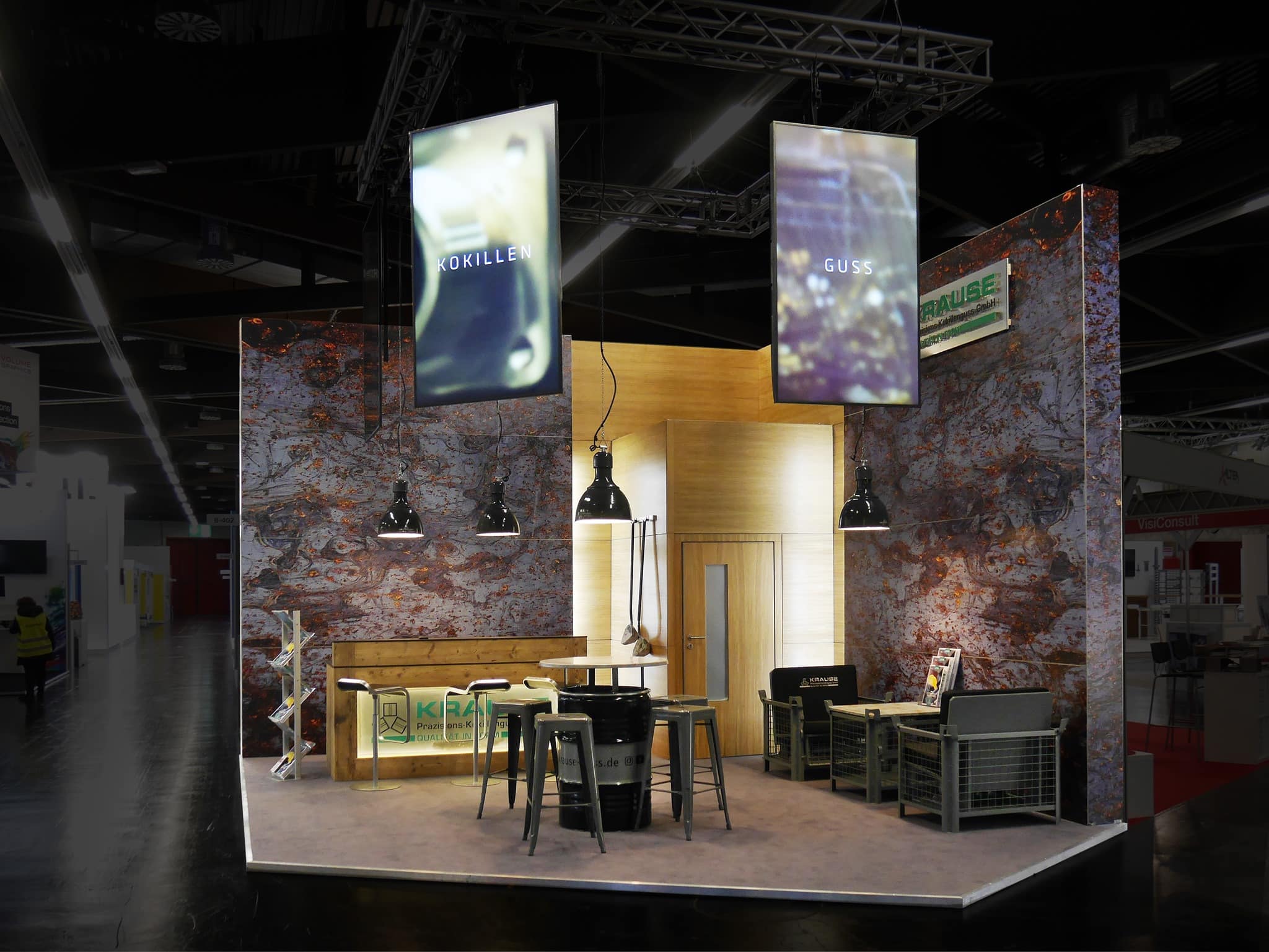 Ein moderner Messestand mit Holzkonstruktion, Industrieleuchten und Metallmöbeln. Darüber hängen zwei große, beleuchtete Schilder. Der Hintergrund ist in einem strukturierten, erdigen Design gehalten. Auf den Auslagetischen liegen mehrere Broschüren aus.
