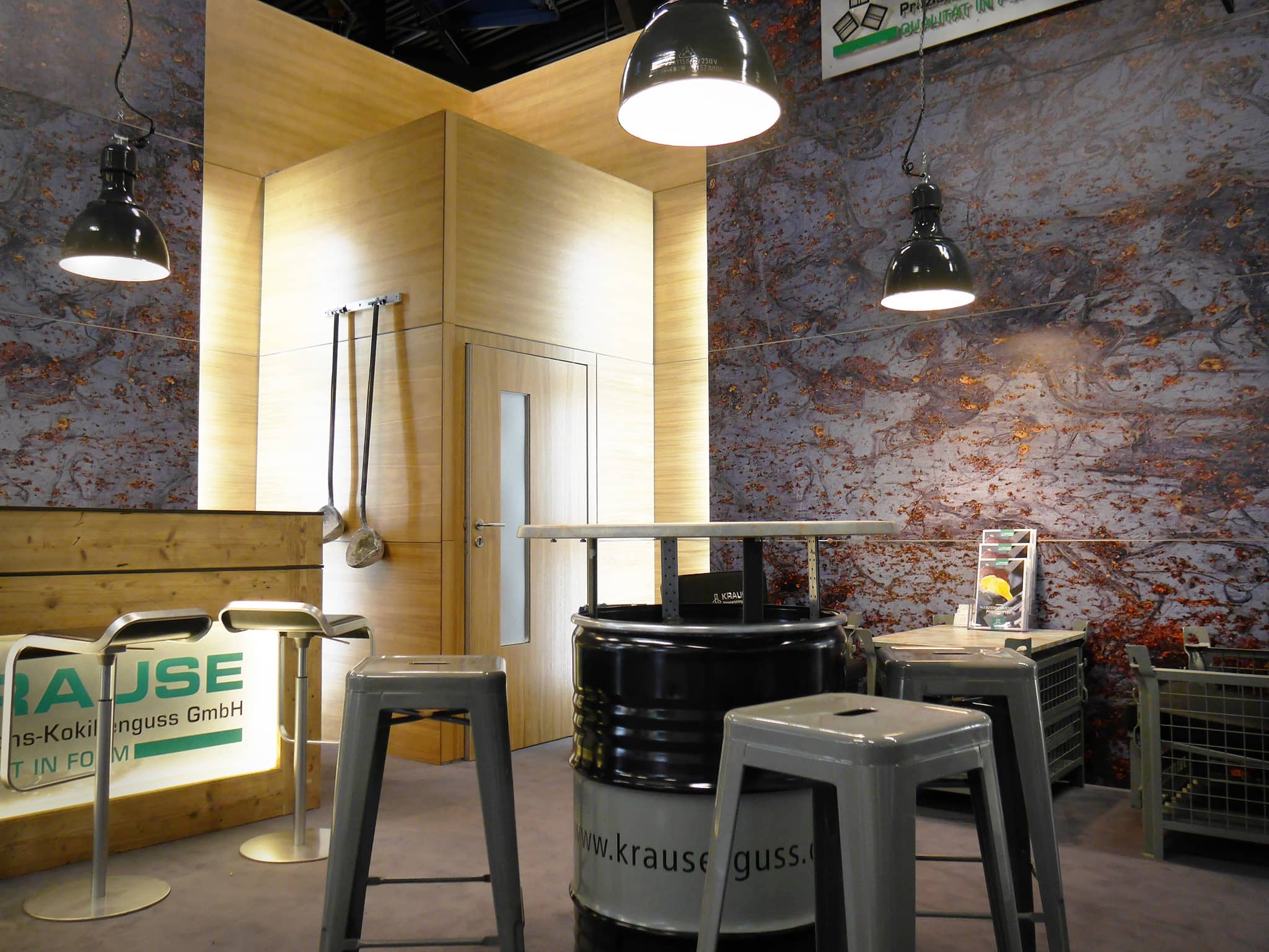 Messestand im Industrie-Stil mit einem improvisierten Tisch aus einem Fass, umgeben von Metallhockern. Der Stand ist mit Holz- und Steintexturen dekoriert, darüber hängen Pendelleuchten, und ein Firmenschild ist gut sichtbar angebracht.