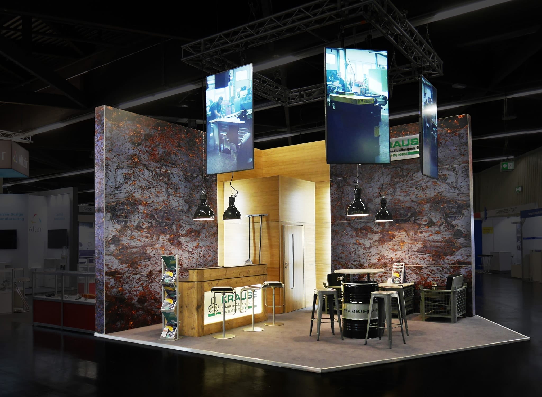 Ein moderner Messestand mit Holz- und Metalldekor verfügt über eine Theke, hohe Hocker und trommelförmige Tische. Überkopfbildschirme zeigen Bilder, und die Wände sind in einem strukturierten, erdigen Design gehalten. Pendelleuchten im Industriestil erhellen den Raum.
