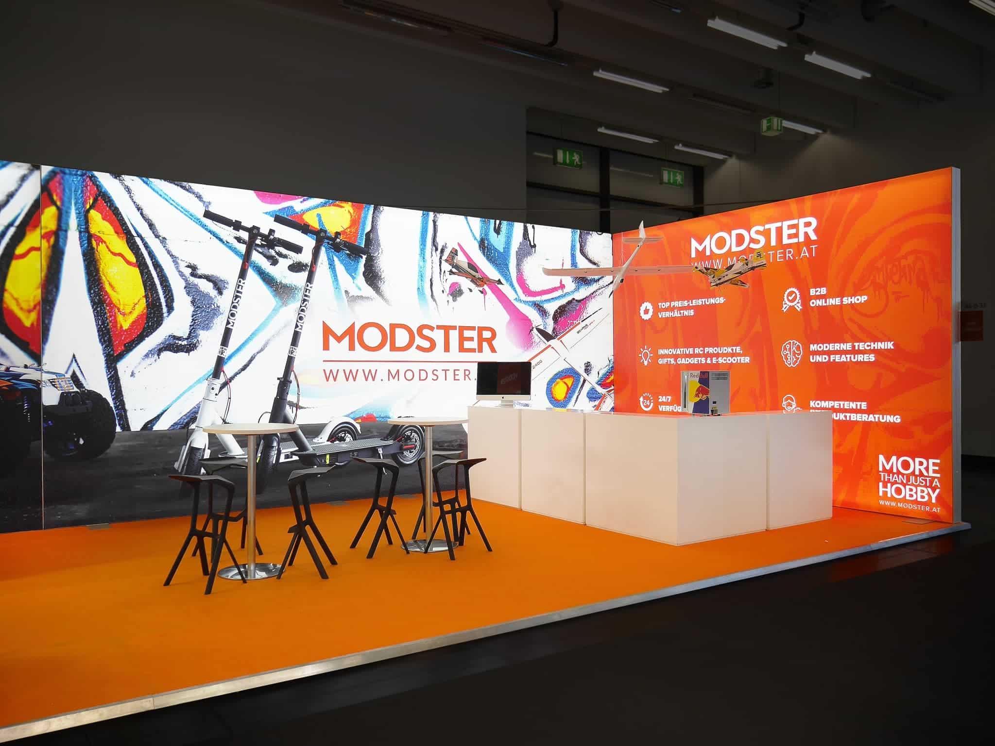 Stand mit Modster-Branding auf einer Messe mit Tischen, Stühlen und farbenfrohen Schautafeln im Graffiti-Stil. Die Tafeln listen Dienstleistungen wie Online-Shopping und technischen Support auf. Der Boden ist leuchtend orange.