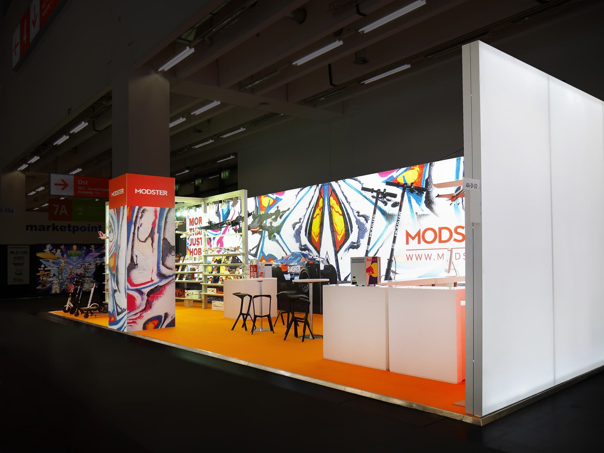 Ein hell erleuchteter Messestand mit dem „Modster“-Branding. Er besticht durch farbenfrohe abstrakte Kunst an den Wänden, eine Theke, Regale mit Produkten und Hocker. Der Boden ist mit orangefarbenem Teppich ausgelegt, und der Stand befindet sich in einer großen Ausstellungshalle.