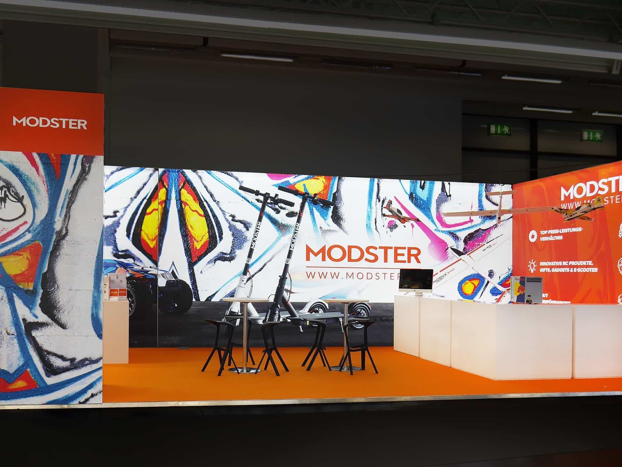 Ein Messestand mit einem farbenfrohen, abstrakten Wandbild mit Schmetterlingen. Der Stand zeigt einen Elektroroller, einen Tisch mit Stühlen und einen Computermonitor. Der Markenname „MODSTER“ ist prominent platziert.