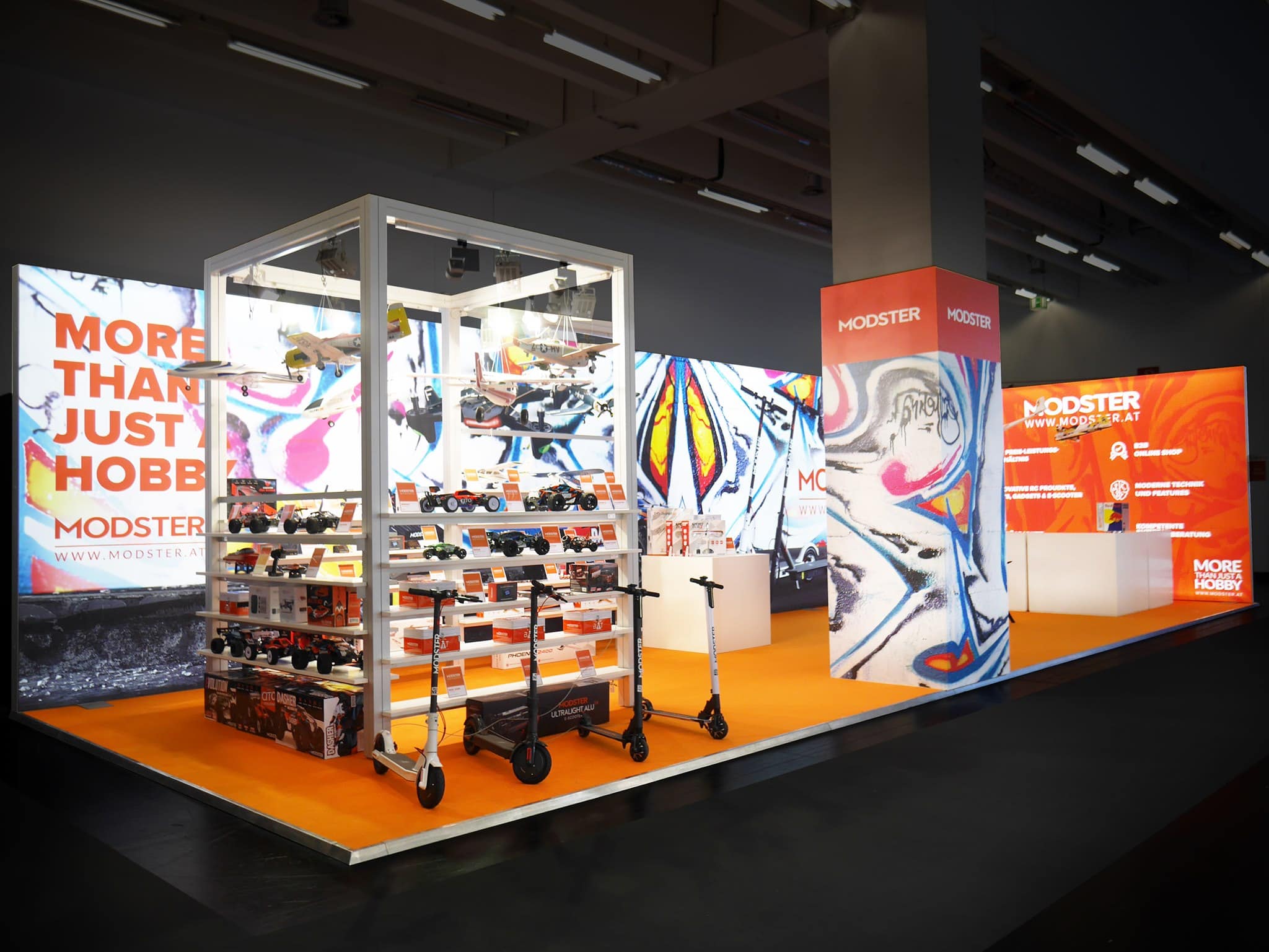 Ein Messestand mit einer Vitrine mit Drohnen und Scootern auf orangefarbenem Boden. Im Hintergrund leuchten hell leuchtende Schilder mit den Aufschriften „Mehr als nur ein Hobby“ und „Modster“. An den Wänden sind bunte, abstrakte Designs zu sehen.