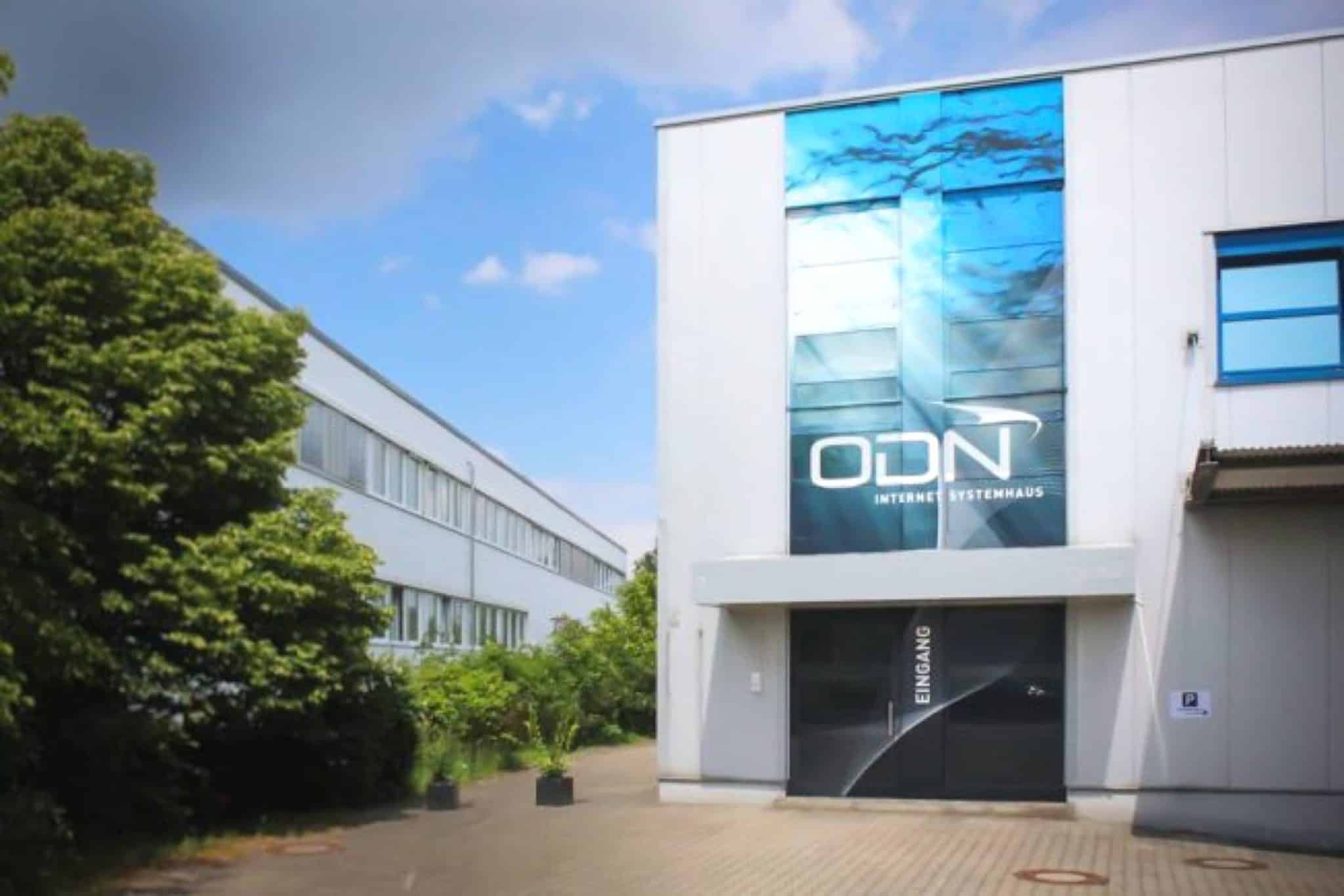 An der Außenseite dieses modernen Gebäudes ziert ein großes vertikales Schild mit der Aufschrift „ODN“ und „Internet Systemhaus“, was auf einen umfangreichen Innenausbau hindeutet. Der Eingang ist über einen von Bäumen und Büschen gesäumten Weg unter leicht bewölktem Himmel erreichbar.