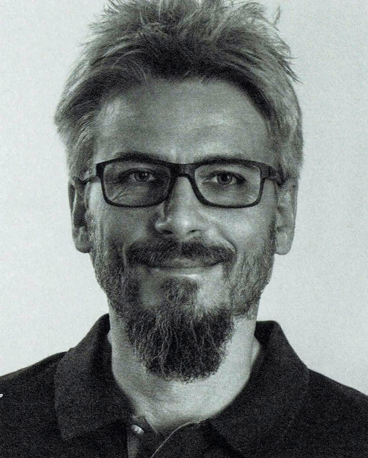 Ein Schwarzweißfoto eines Mannes mit kurzen Haaren, Brille und Bart. Er lächelt leicht und trägt ein dunkles Hemd mit Kragen vor einem schlichten Hintergrund.