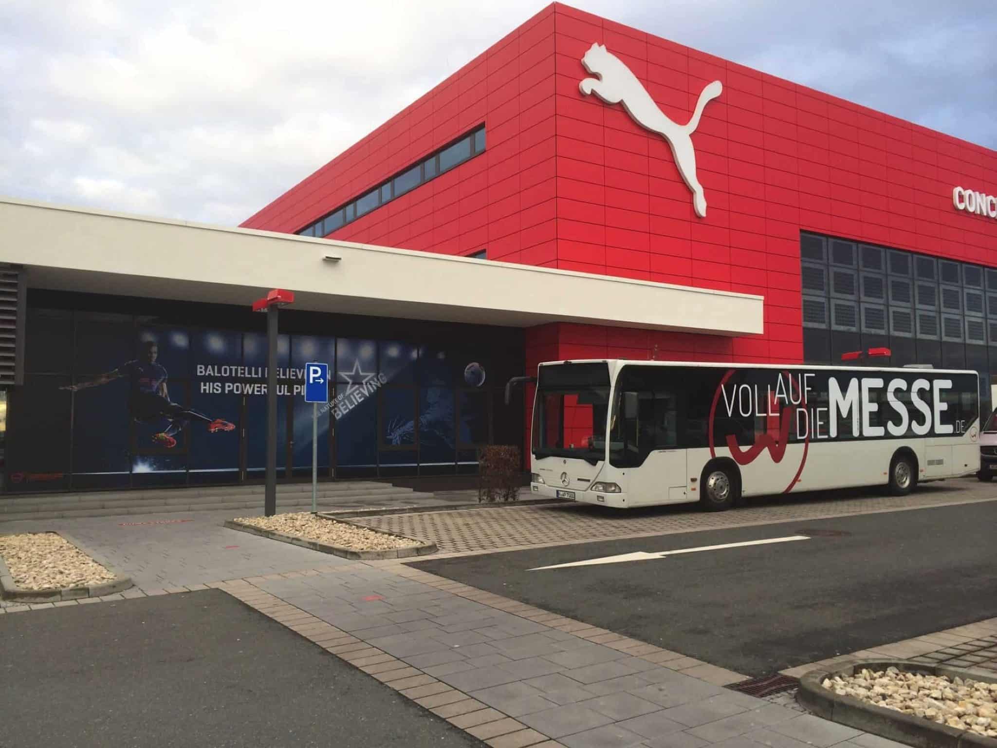 Ein Bus mit der Aufschrift „VOLL AUF DIE MESSE“ parkt vor einem leuchtend roten Puma-Gebäude und präsentiert dessen beeindruckende Innenausstattung. Die Fassade ist mit großen Puma-Logos und Fußball-Grafiken verziert, während der Parkplatz und der gepflasterte Gehweg das lebendige Bild abrunden.