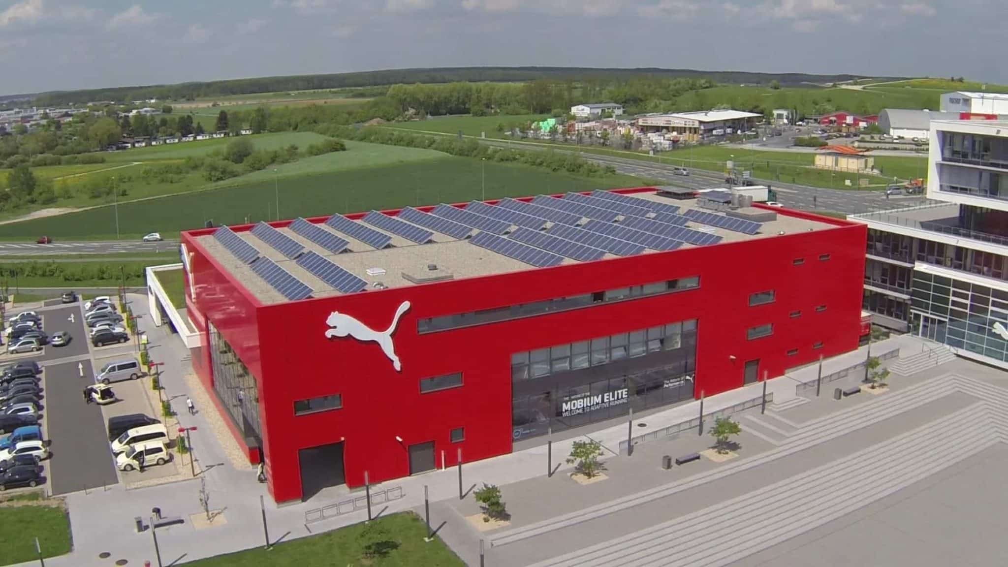 Ein großes rotes Gebäude mit Solarmodulen auf dem Dach und einem weißen Puma-Logo an der Seite erinnert an das moderne Design von Referenzen Messebau. Umgeben von Parkplätzen und grünen Feldern thront es stolz unter einem klaren, sonnigen Himmel.