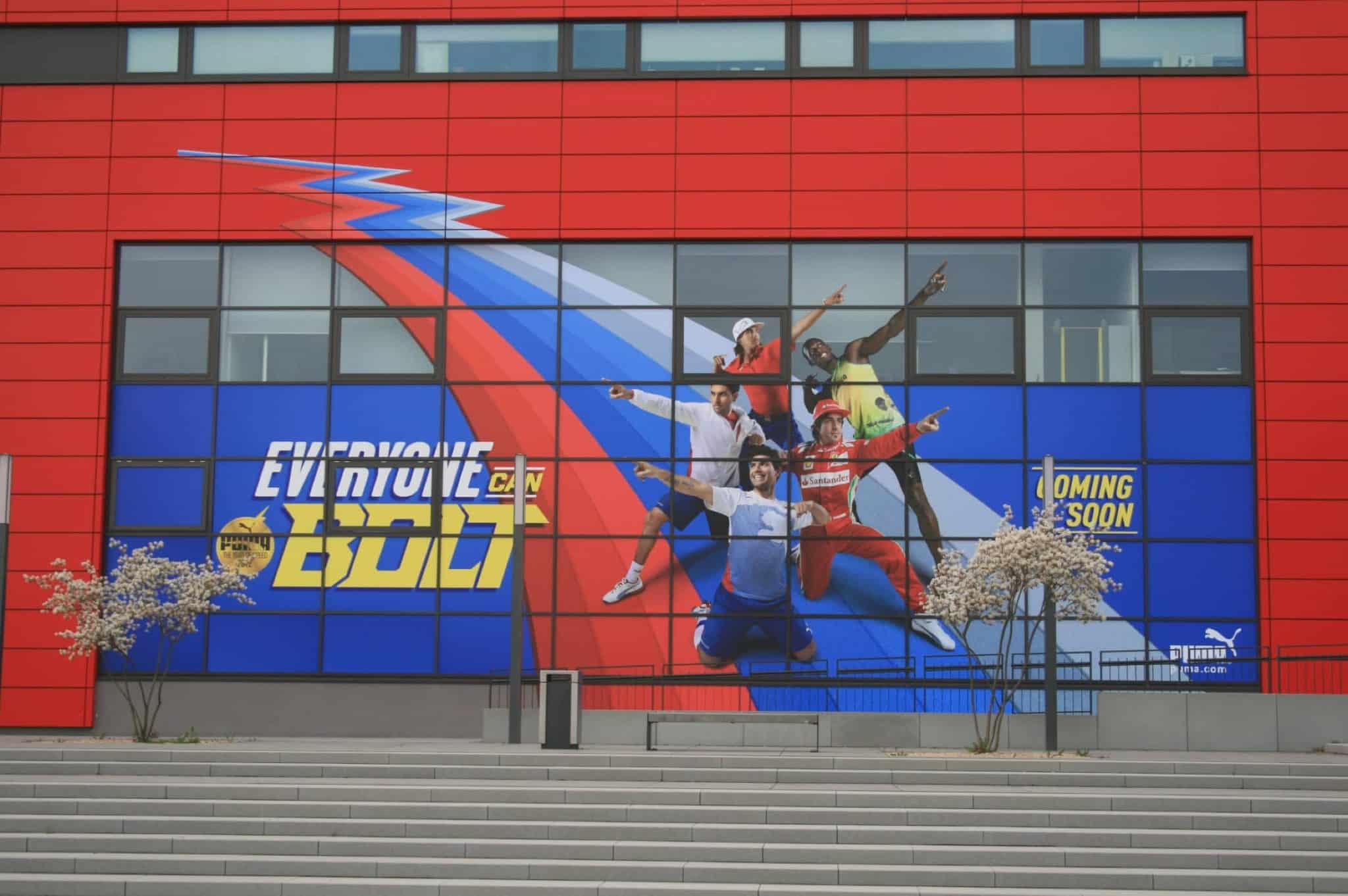 Ein großes Gebäude präsentiert eine farbenfrohe Werbung in den Farben Blau, Rot und Weiß. Sie zeigt Sportler in dynamischen Posen. Der Text lautet „Everyone on Bolt“ und „Coming Soon“ neben dem Puma-Logo. Im Hintergrund sind laufende Innenausbauarbeiten zu sehen, die eine spannende Enthüllung versprechen.