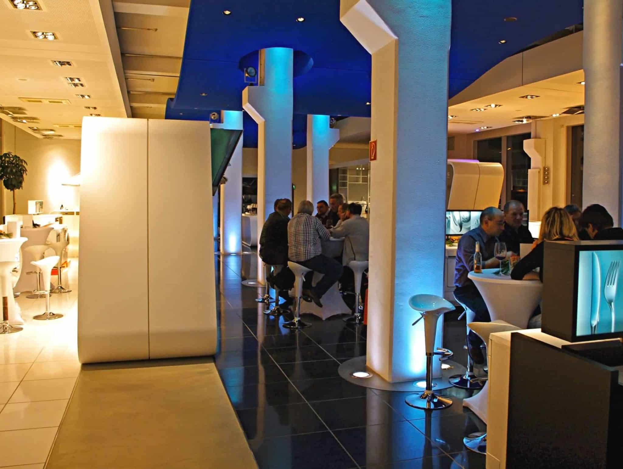 Ein modernes Restaurant mit blauem Ambientelicht und weißen Möbeln bietet Platz für lebhafte Veranstaltungen. Die Gäste sitzen an einem langen Tisch, essen und unterhalten sich. Hohe weiße Säulen und Barhocker unterstreichen die elegante, moderne Atmosphäre.