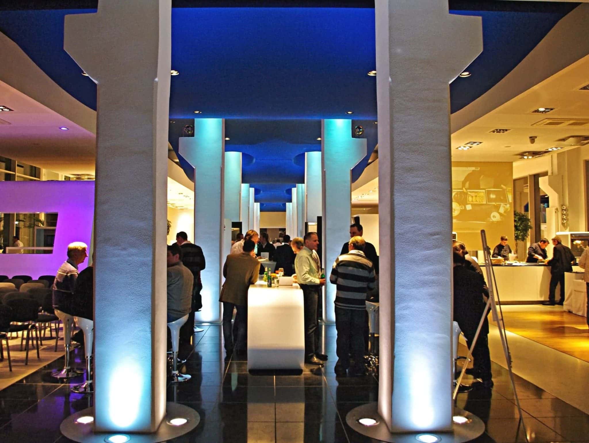 Eine moderne Bar mit hohen weißen Säulen und blauem Licht bietet lebhafte Veranstaltungen. Gruppen von Gästen versammeln sich um Stehtische und unterhalten sich. Die Atmosphäre ist geprägt von einem Mix aus hellem Weiß und sanftem Lila.