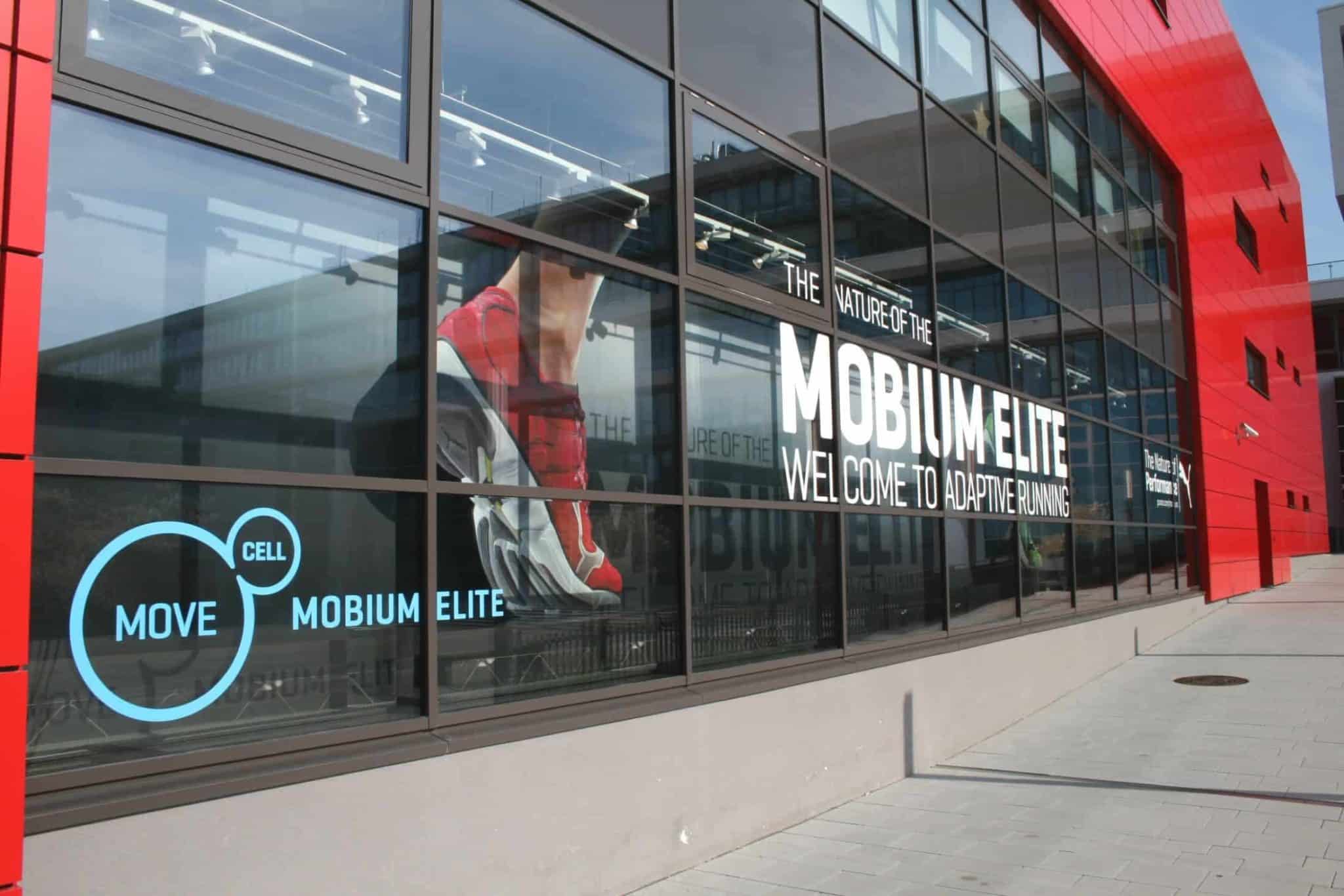 Die Glasfassade des Gebäudes präsentiert eine auffällige Werbung mit einem Läufer in roten Schuhen. Der Text verkündet: „Die Natur des Mobium Elite: Willkommen beim adaptiven Laufen“, mit Grafiken und kleinerem Text „Move Cell Mobium Elite“. Rote Strukturdetails unterstreichen das innovative Innenausbau-Design.