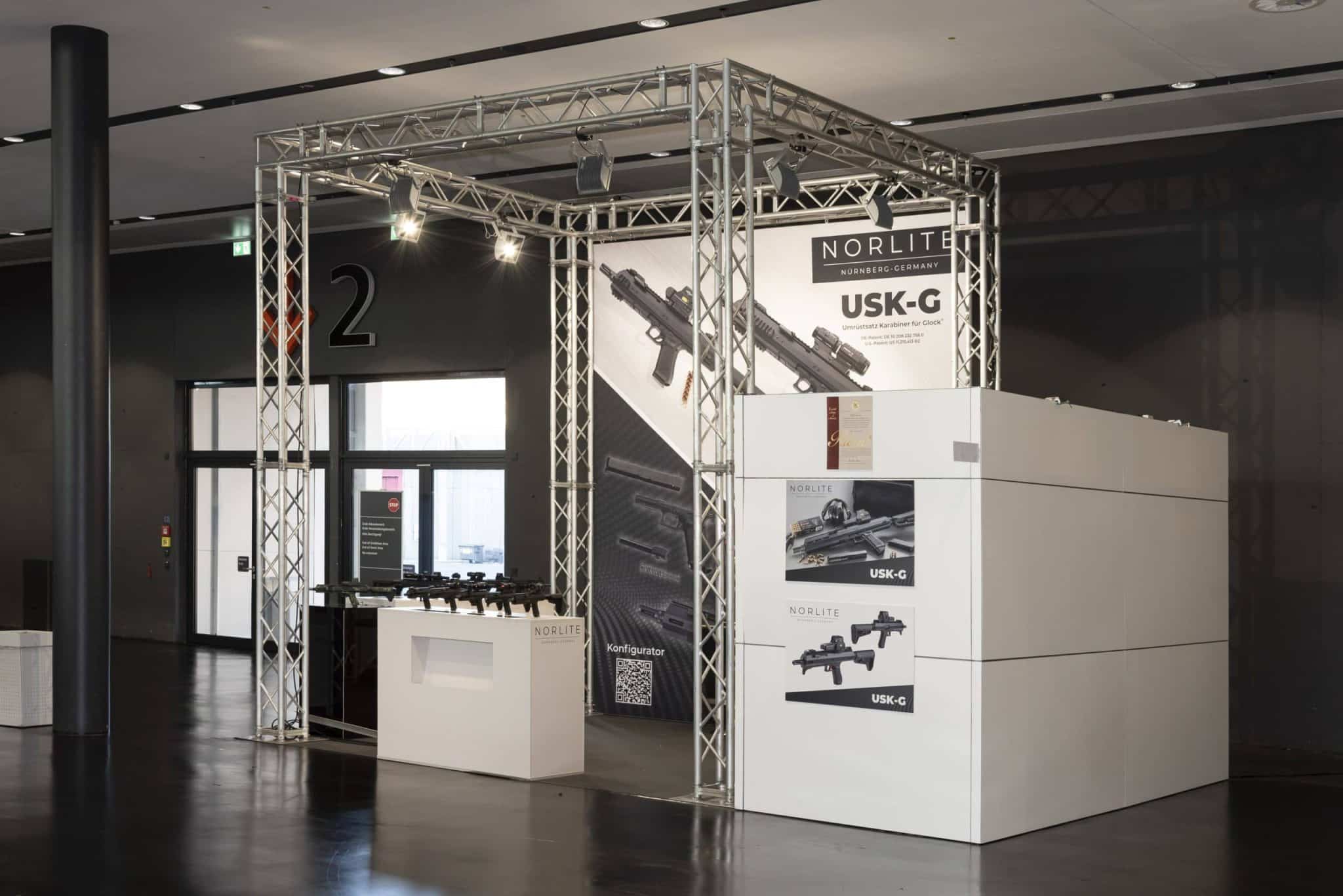 Messestand von Norlite auf der IWA OutdoorClassics in Nürnberg mit modernem Standbau und großem Banner der USK-G Waffenserie.