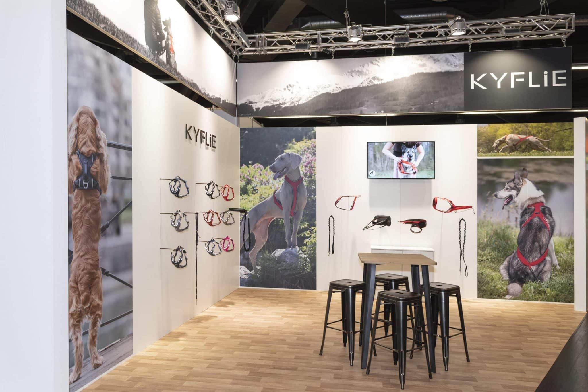 Helle Messestandgestaltung von Kyflie auf der Interzoo Nürnberg mit Wandbildern von Hunden, ausgestellten Hundegeschirren und Stehtisch im Zentrum.