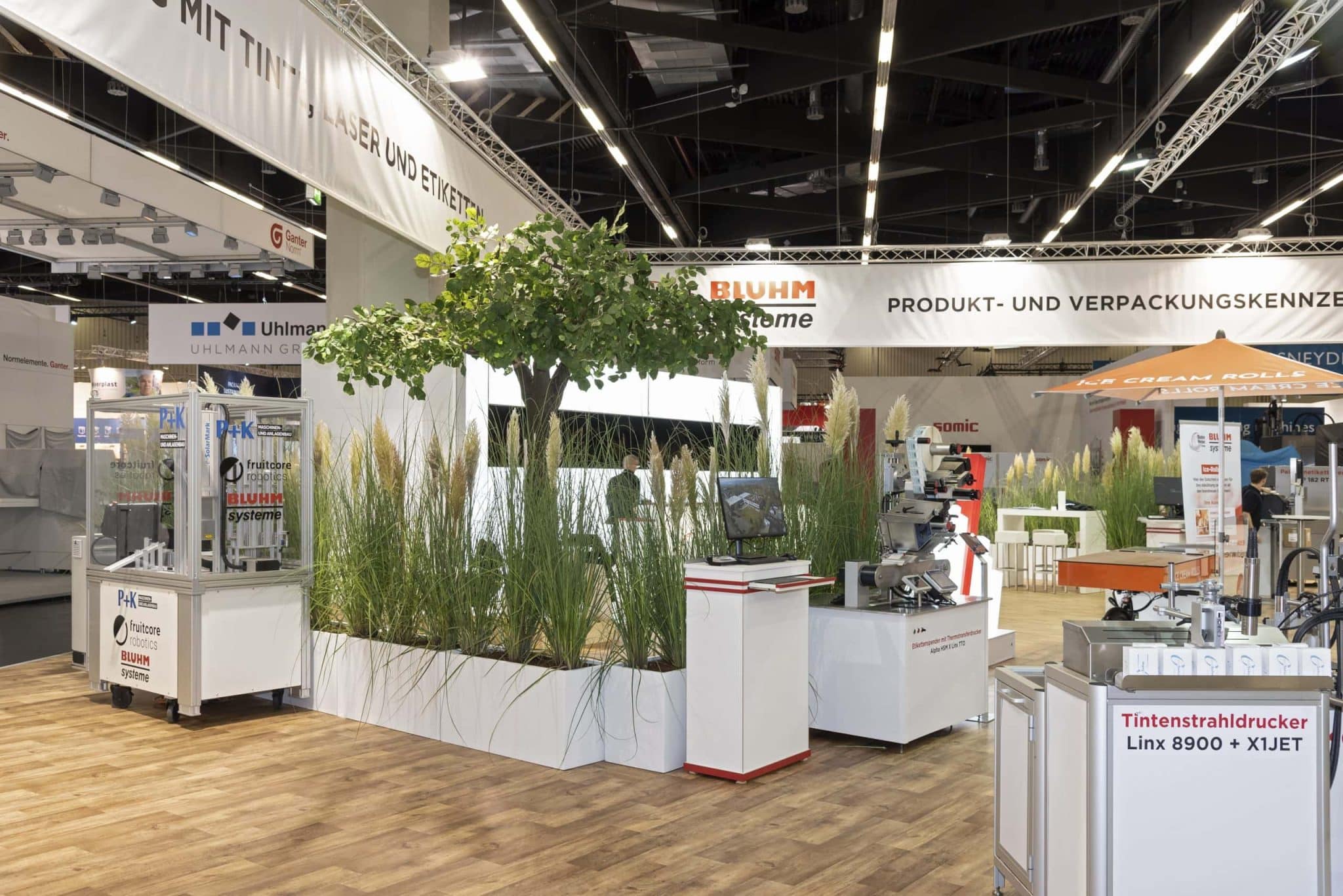 Bluhm Systeme Messestand mit Robotik und Tintenstrahldruckern, eingebettet in grünes Messebau-Design – Messebau Nürnberg.