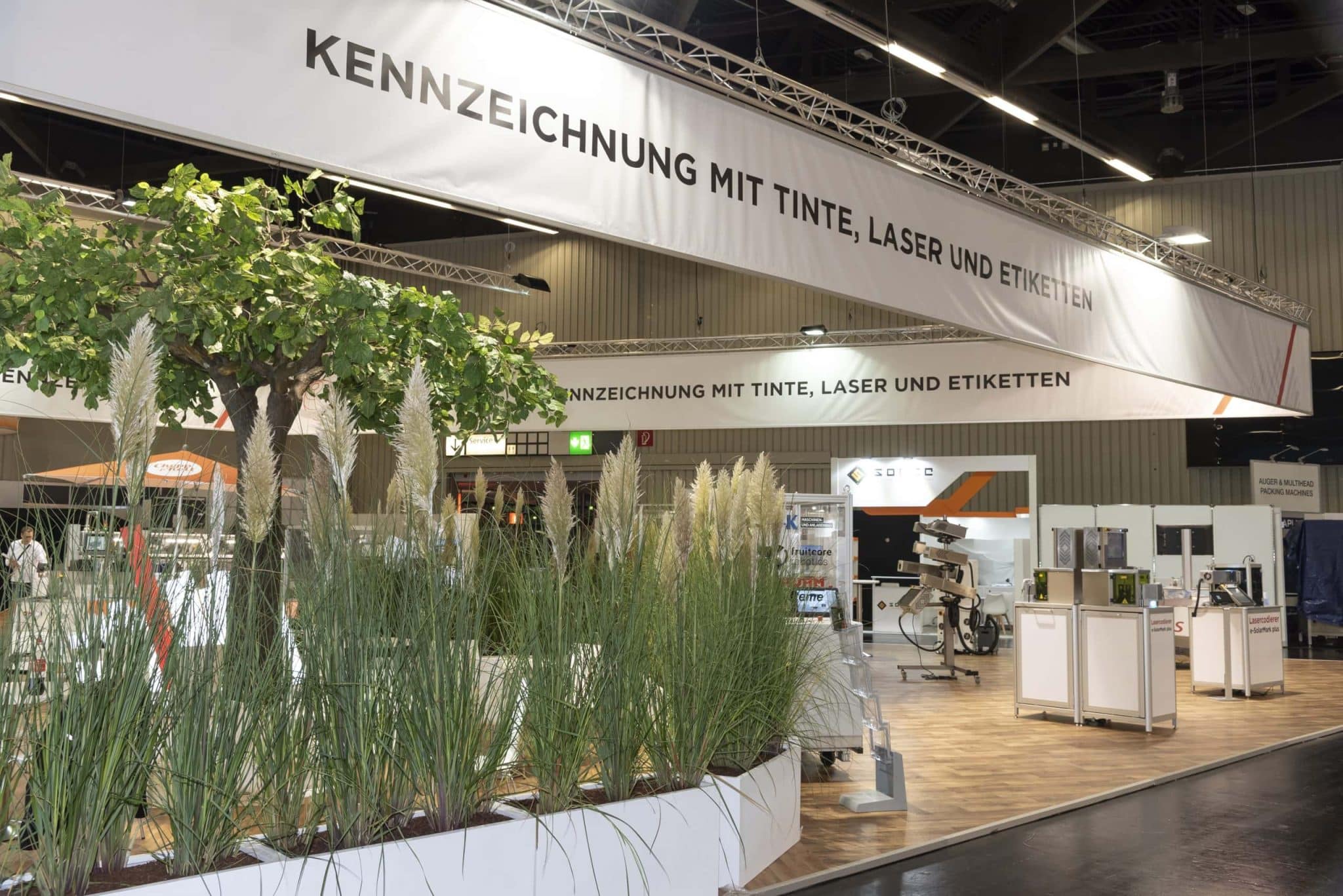 Begrünter Messestand mit Großbanner „Kennzeichnung mit Tinte, Laser und Etiketten“ von Bluhm Systeme – Messebau Nürnberg.