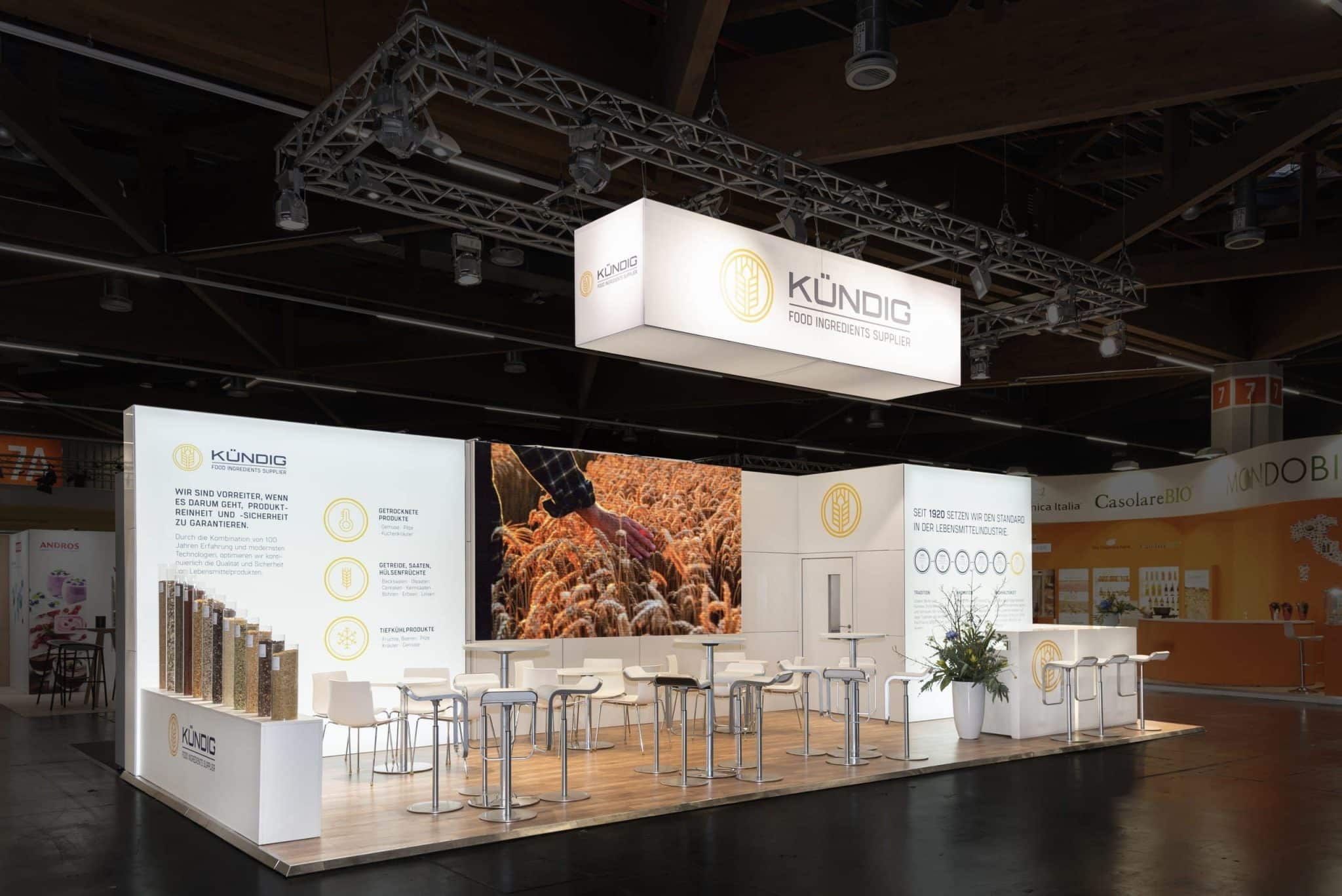 Moderner Messestand von Kündig auf der Biofach 2025 in Nürnberg mit beleuchtetem Logo, Infowänden und Barhockern im Vordergrund.