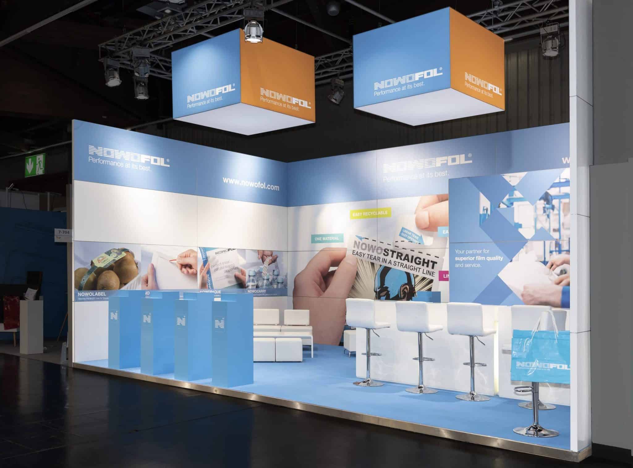 Fachpack Nürnberg Messestand von Nowofol mit weißen Barhockern, Sitzbereich und Deckenwürfeln „Messebau Nürnberg“.