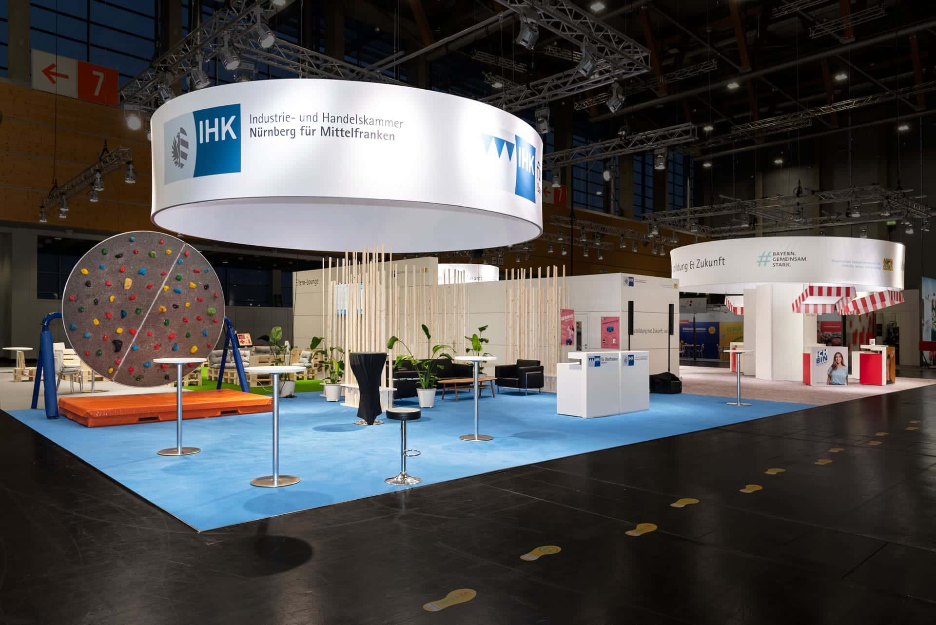 Ein hell erleuchteter Messestand mit kreisförmiger Beschilderung für die IHK Nürnberg für Mittelfranken. Zu den Besonderheiten gehören ein großes buntes Spinnrad, Tische, Stühle, Pflanzen und Informationstafeln auf einem blauen Teppich. Die umliegenden Stände sind sichtbar.