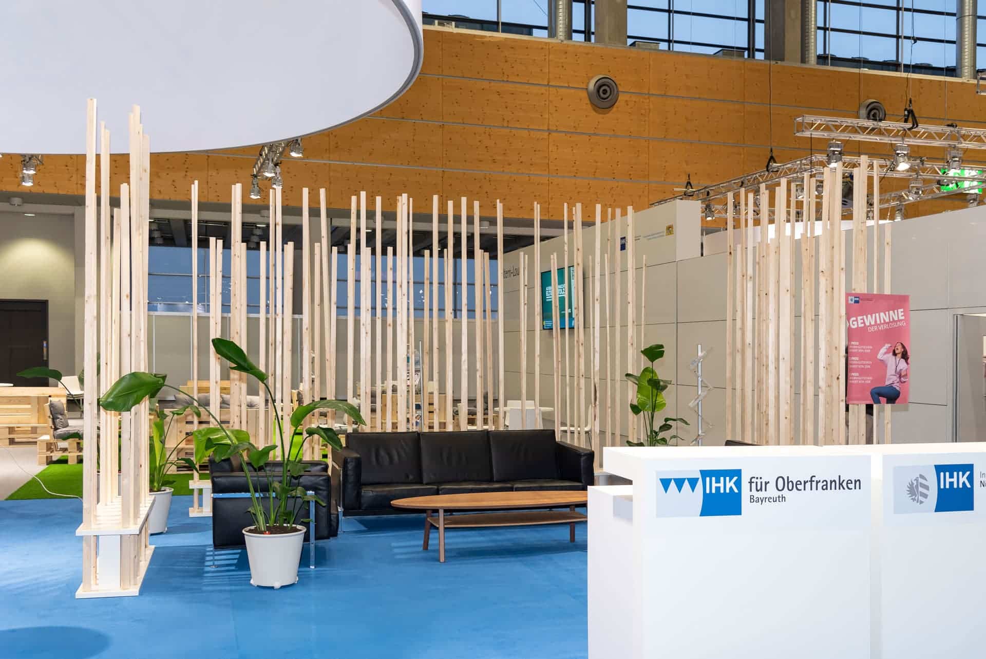 Ein moderner, offener Messestand mit schwarzem Sofa, Holztrennwänden und Topfpflanzen. Der Stand ist mit blauem Teppichboden und Beschilderungen mit Logos und Texten in deutscher Sprache ausgestattet. Helle Beleuchtung und ein Holzhintergrund runden das Ambiente ab.