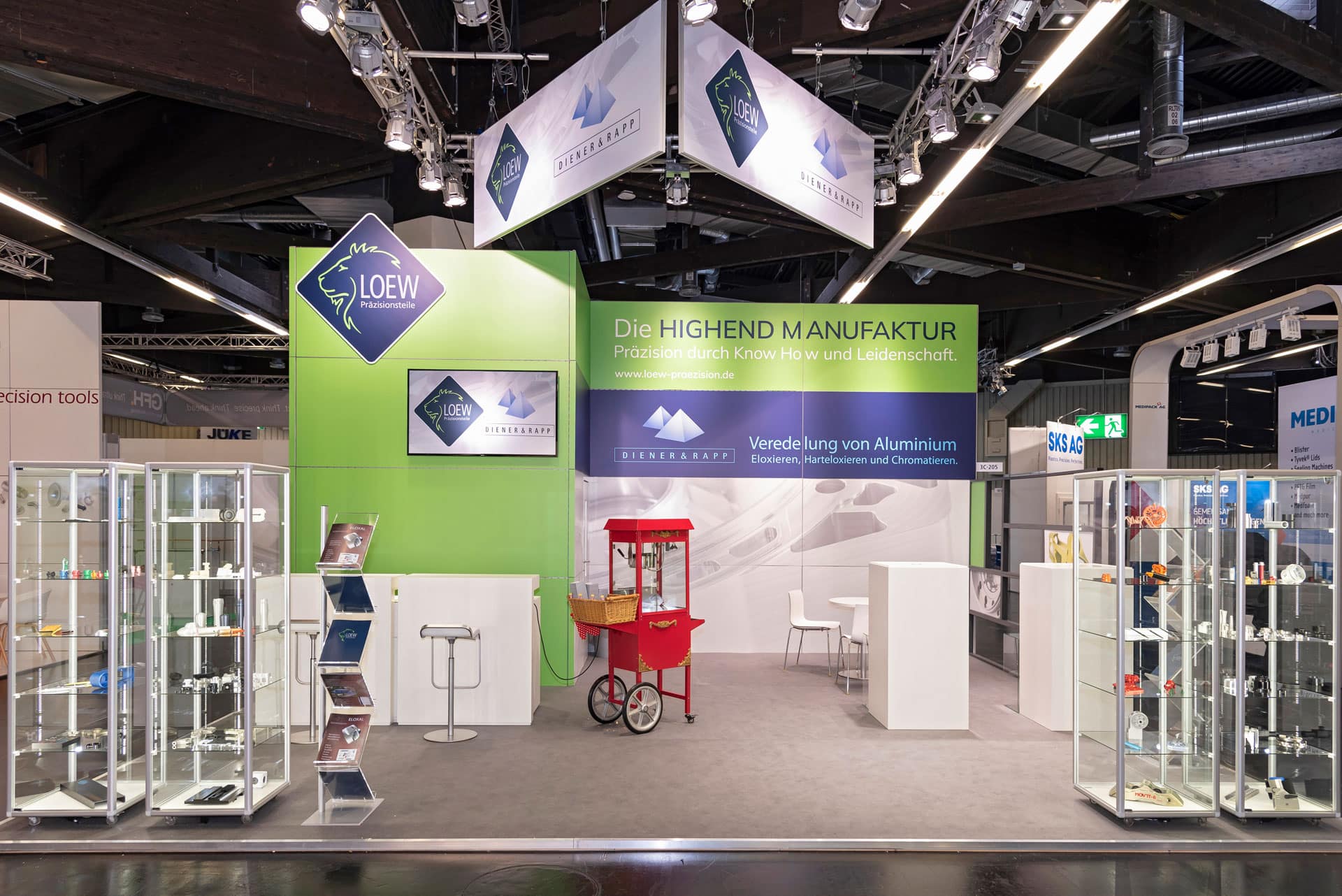 Vorderansicht des Löew Präzisionsteile Messestands auf der Medtech Live 2023 mit Glasvitrinen, roten Popcornwagen und Ausstellungsstücken aus Aluminium.
