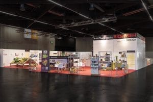 Ein Messestand mit transparenten Glaswänden, einem roten Teppich und verschiedenen Produktpräsentationen. Schilder mit den Aufschriften „Meetingpoint“ und „BIOFACH“ stehen bereit. Im Inneren gibt es Produktregale, Tische und Werbematerialien.
