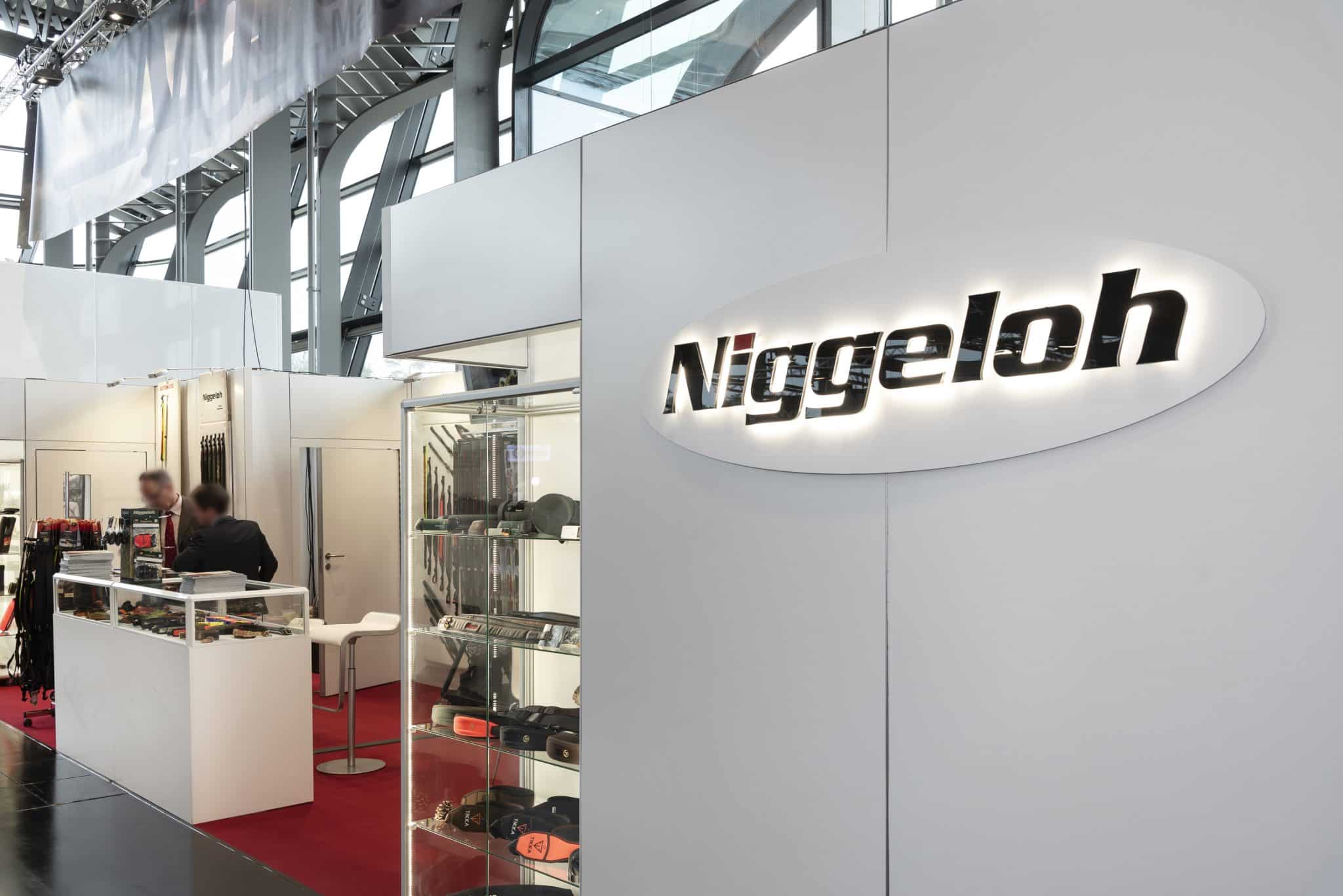 Moderner Messestand von Niggeloh auf der IWA in Nürnberg, mit Glasvitrinen, beleuchtetem Logo und rotem Teppich – Messebau Nürnberg