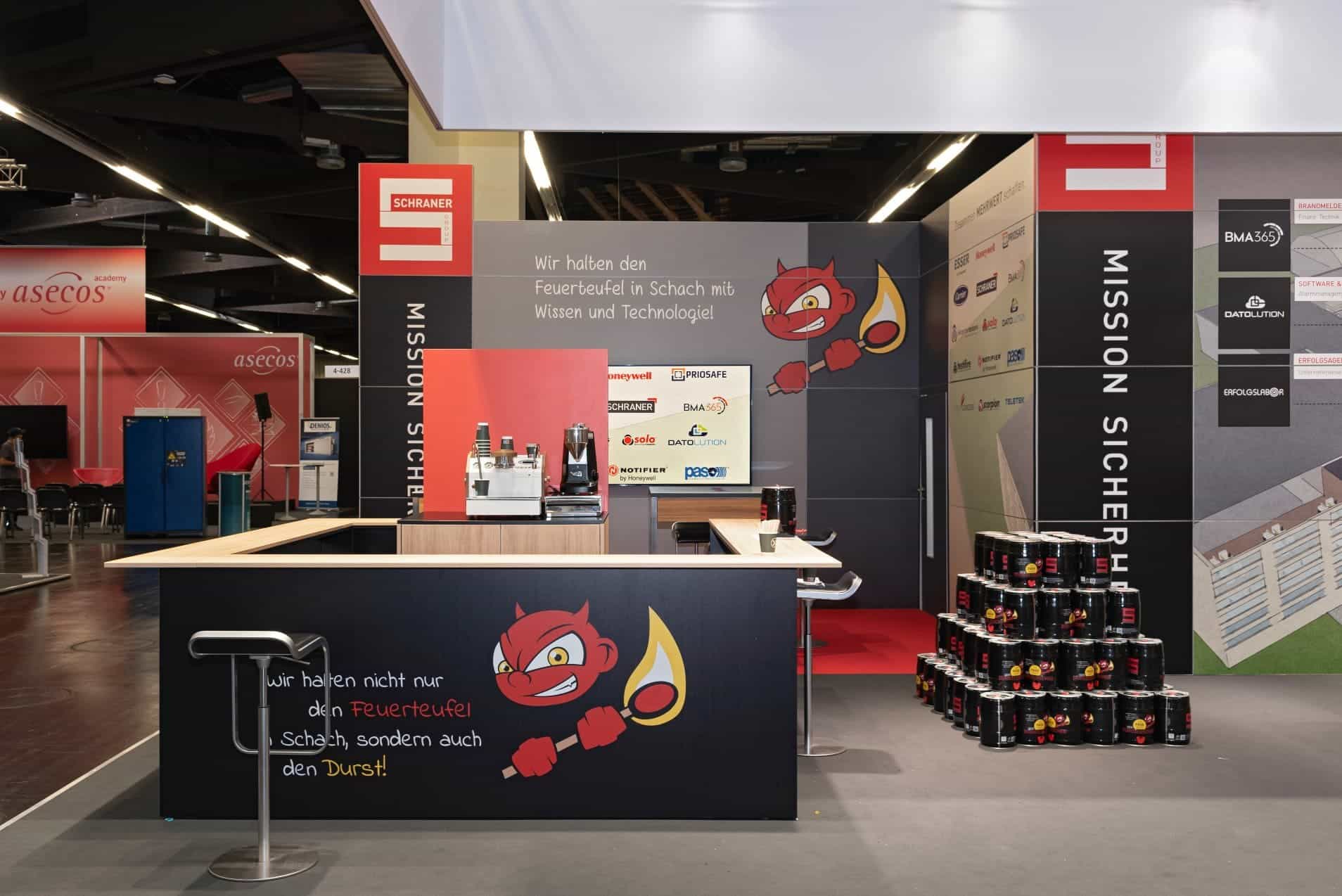 Messestand von Schraner Group auf der FeuerTrutz in Nürnberg mit Theke, Kaffeemaschine und Feuerteufel-Maskottchen.