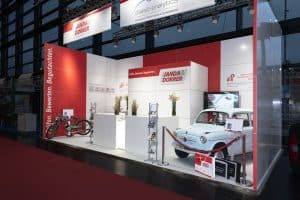Besuchen Sie unseren Messestand auf der Automobilmesse mit einem blauen Oldtimer und einem klassischen Fahrrad. Der rot-weiße Stand ist mit Informationsplakaten, Broschüren und Blumenarrangements geschmückt. Ein roter Teppich führt Sie elegant in dieses nostalgische Automobilerlebnis.