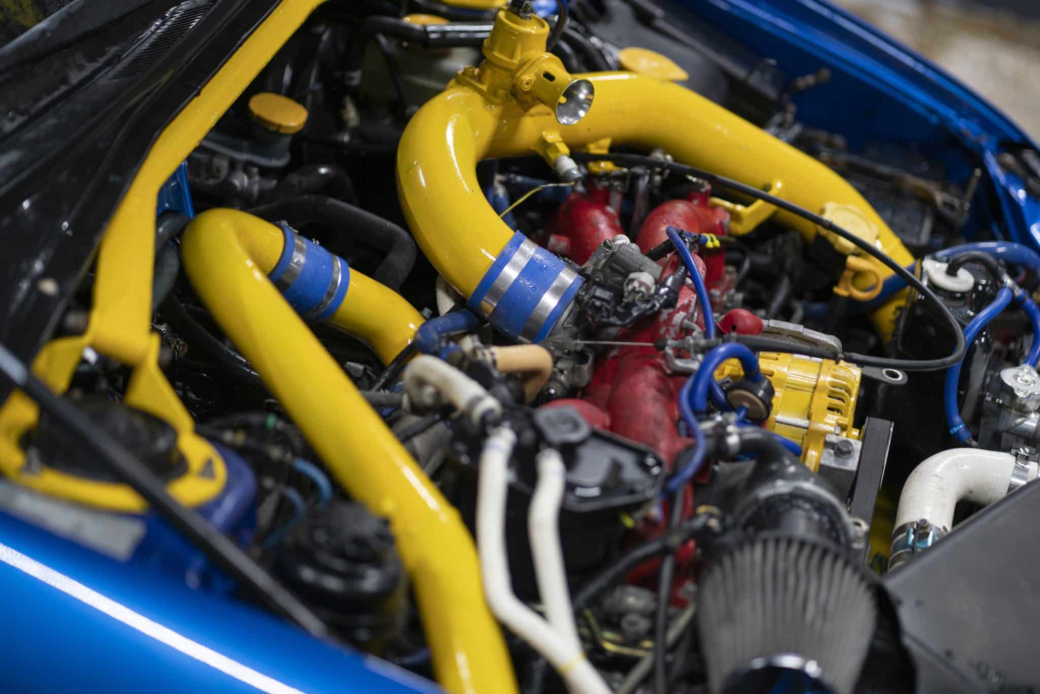 Nahaufnahme eines Automotors mit leuchtend gelben Schläuchen und Komponenten, perfekt für die Präsentation auf der Automobilmessen. Der Motor besticht durch einen roten Ansaugkrümmer und blaue Akzente, die mit verschiedenen Kabeln und Schläuchen verflochten sind. Die blaue Außenfarbe des Autos ist rund um den Motorraum sichtbar und unterstreicht sein Design.