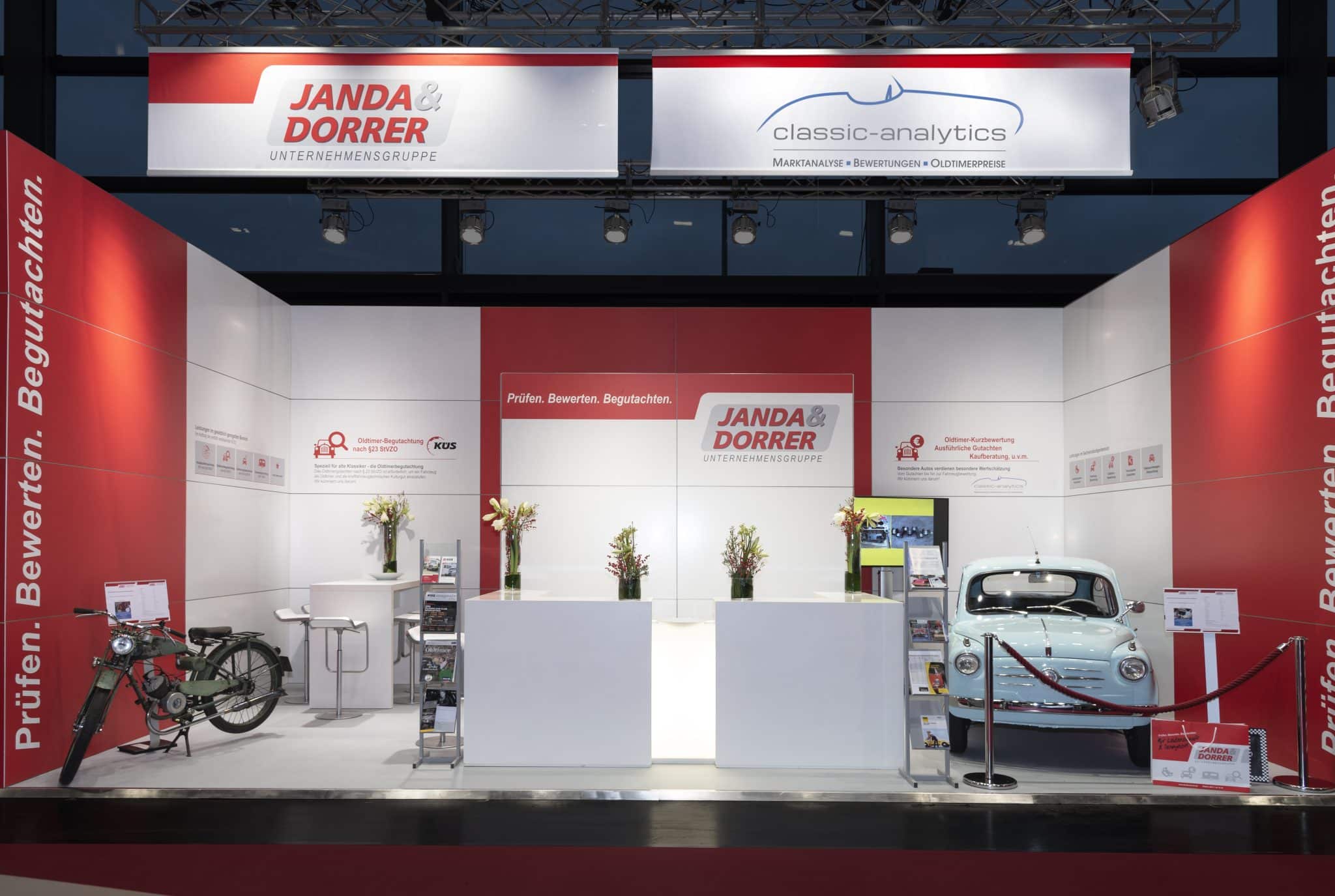 Der Messestand von Janda & Dorner auf der Automobilmessen besticht durch sein markantes rot-weißes Farbkonzept. Ein klassisches Motorrad und ein Oldtimer harmonieren perfekt mit modernen Möbeln und Blumenarrangements. Die Beschilderung hebt die Bewertungs- und Prüfdienstleistungen hervor.