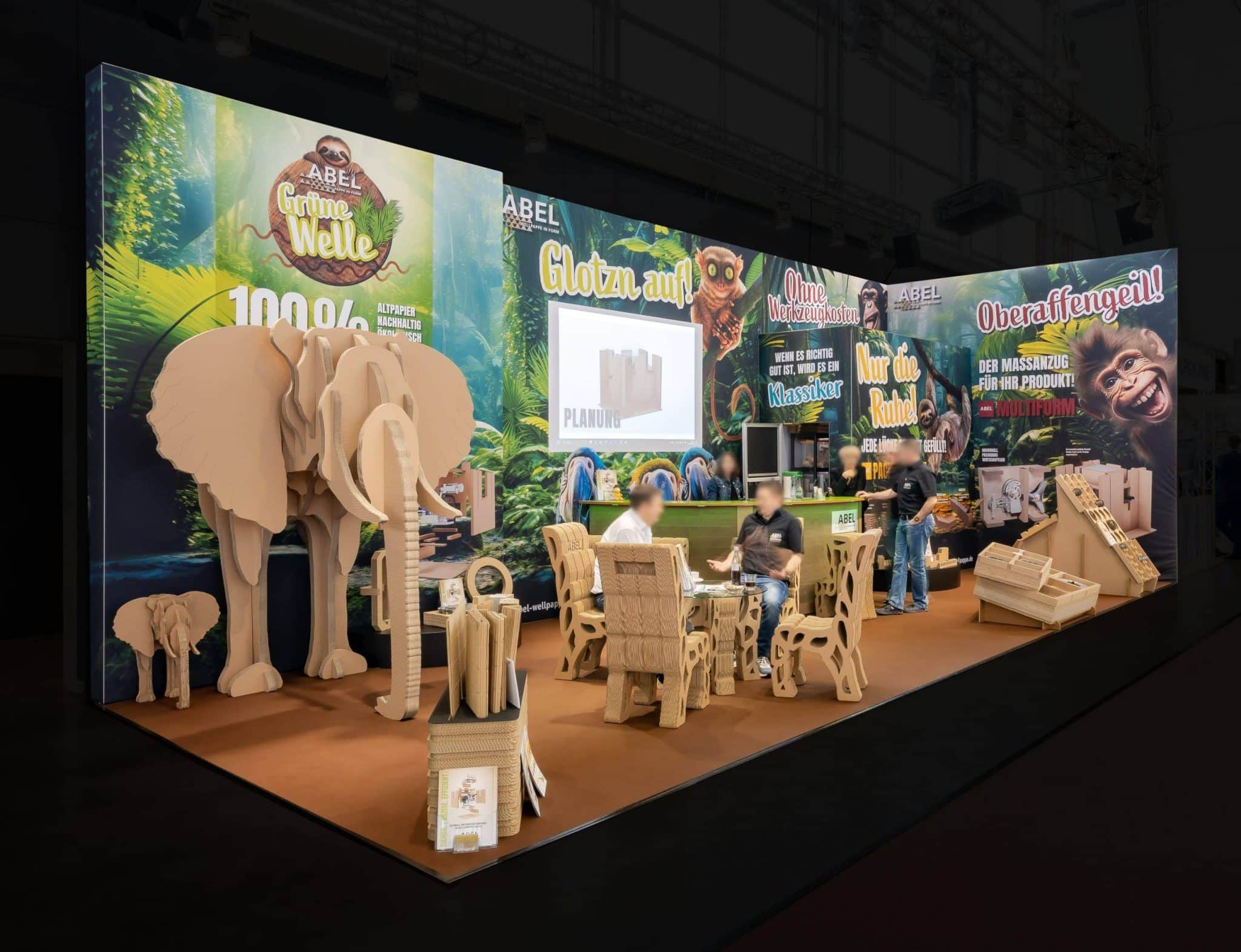 Auf dem größten Messegelände Deutschlands präsentiert unser Messestand innovative Pappmöbel und -dekorationen, darunter Elefanten, Stühle und Tische. Das Dschungel-Setup besticht durch leuchtend grüne Hintergründe und farbenfrohe Banner und lädt Besucher zum fantasievollen Betrachten ein.