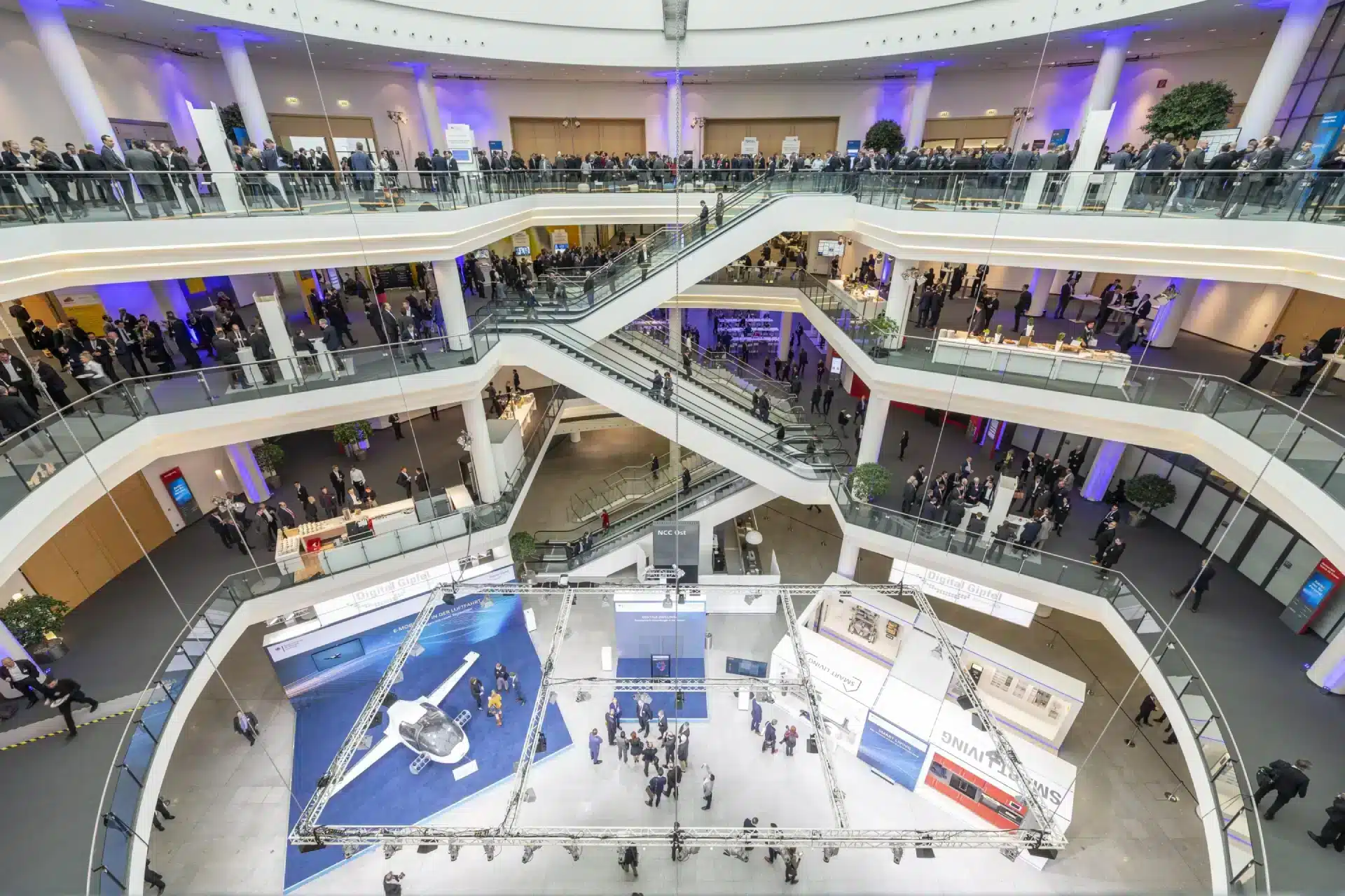 Im Inneren des großzügigen, mehrstöckigen Kongresszentrums der NürnbergMesse herrscht auf mehreren Etagen reges Treiben. Besucher können sich in der Nähe der Ausstellungen versammeln und sich über Rolltreppen und Glasgeländer bewegen. Zahlreiche Messestände und Leuchtdisplays beleben den weitläufigen Raum.
