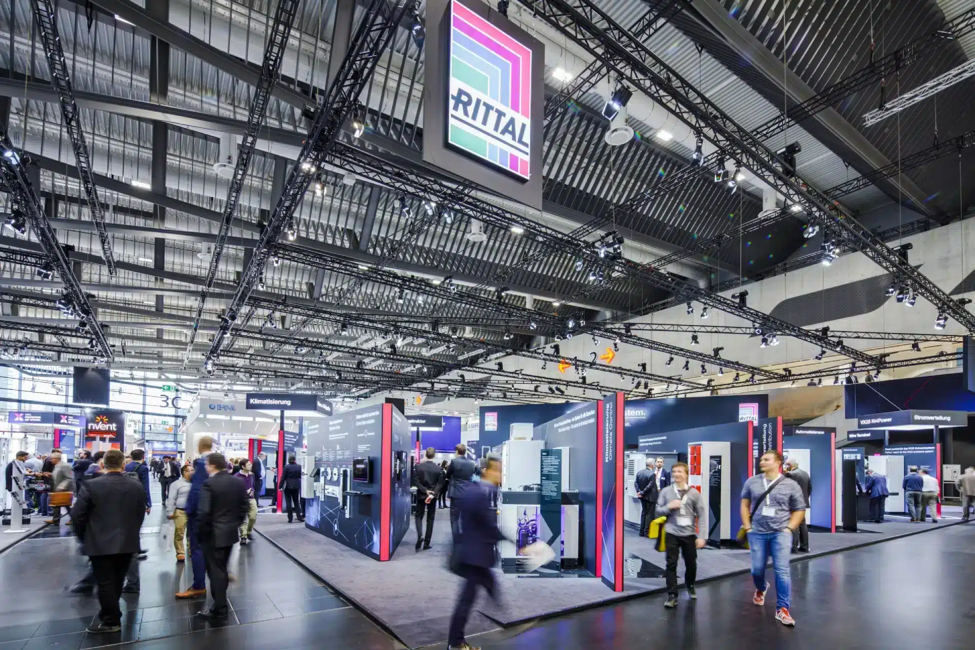 Besucher schreiten durch eine große, moderne Messehalle der NürnbergMesse, umgeben von verschiedenen Unternehmensständen. Die Decke ist mit einem komplexen Lichtnetz versehen, ein markantes Schild trägt den Namen „RITTAL“. Zwischen farbenfrohen Ausstellungen treten Aussteller mit Besuchern in Kontakt.