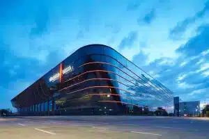 Ein modernes Gebäude mit geschwungener Architektur und Glasfassade, die mit horizontalen roten Lichtern geschmückt ist, steht unter einem bewölkten, blauen Himmel. Die Struktur spiegelt sich deutlich im Glas und erinnert an das elegante Design der NürnbergMesse, mit einer leeren Straße im Vordergrund.