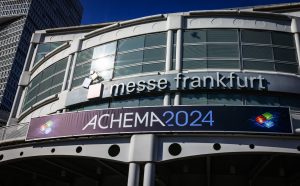 Außenansicht der Messe Frankfurt mit einem prominent platzierten ACHEMA 2024-Banner. Die moderne Architektur des Gebäudes, akzentuiert durch große Fenster, glänzt im Sonnenlicht, während in der Ferne ein Hochhaus aufragt.