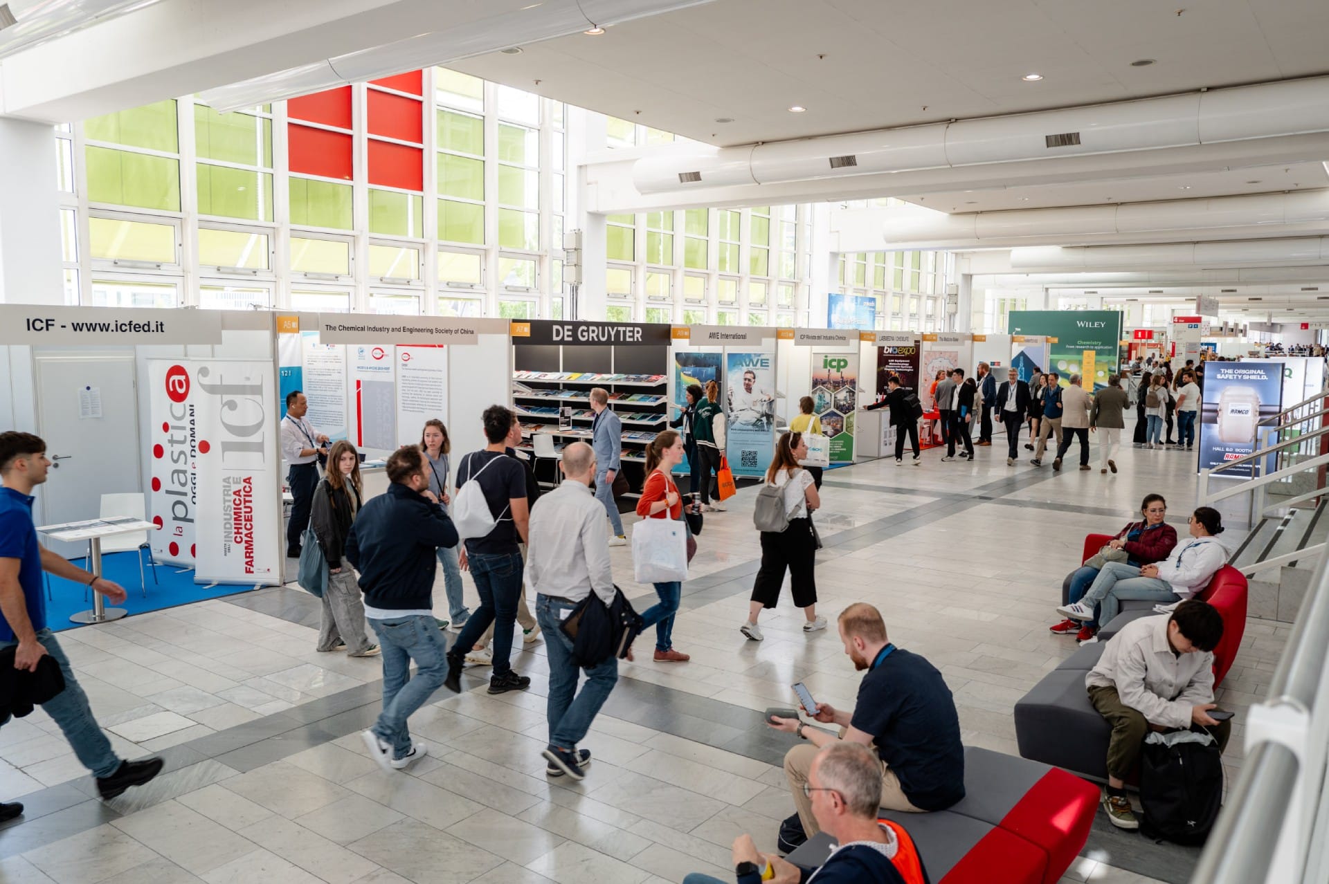 Im geschäftigen Kongresszentrum der Achema herrscht reges Treiben an den Ständen der Aussteller. Der hell erleuchtete Raum mit weißen Wänden und großen Fenstern ist mit Firmenschildern und Bannern geschmückt. Rechts sitzen einige Personen auf roten Bänken und genießen die lebhafte Atmosphäre.