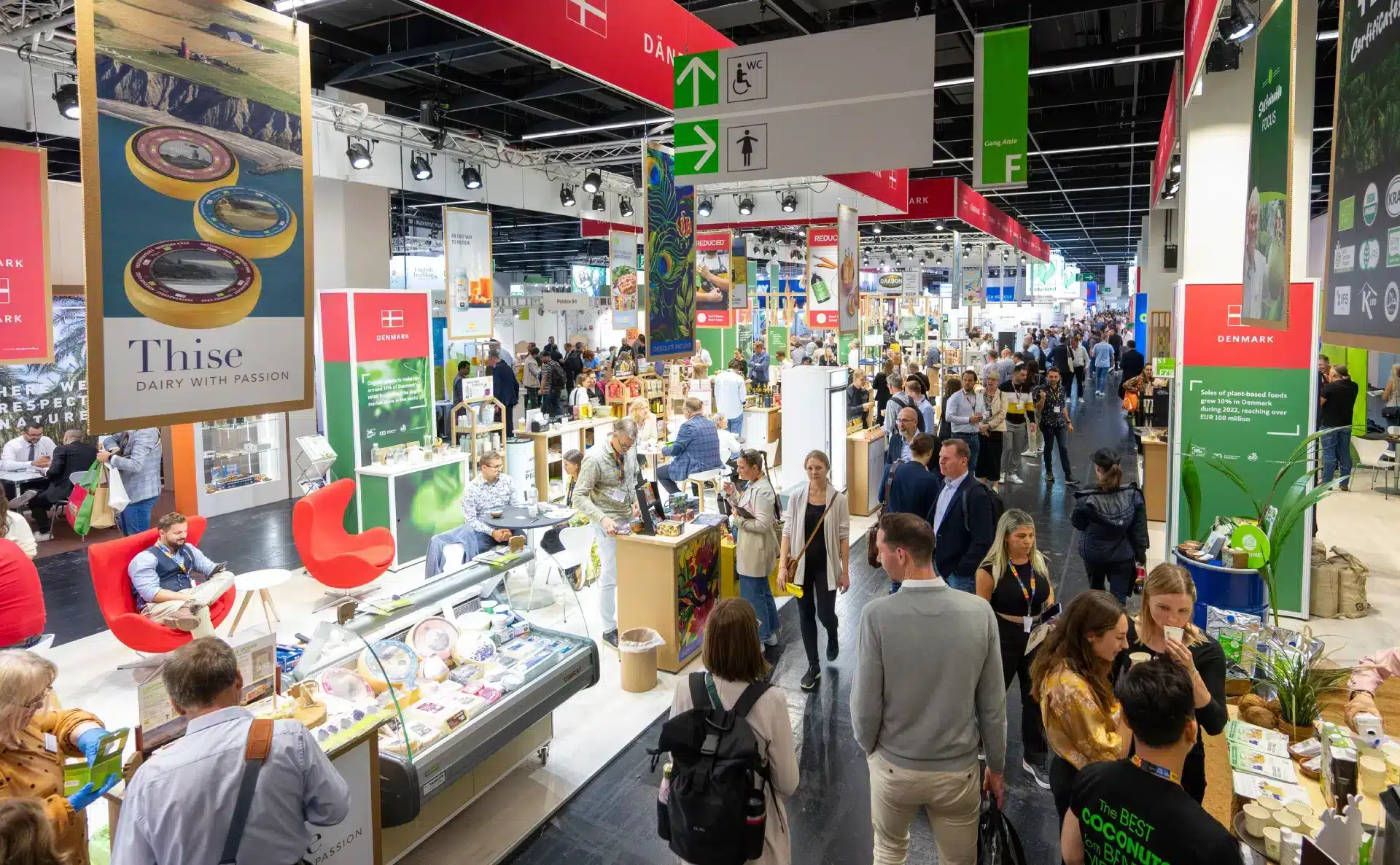 Eine geschäftige Messe, die an die Anuga erinnert, mit einem Austausch zwischen den verschiedenen Ständen, an denen Produkte präsentiert werden. Die Messestände präsentieren farbenfrohe Marken- und Werbematerialien. Überall auf dem Gelände sind Schilder und Deckenleuchten zu sehen, die eine lebendige Atmosphäre schaffen.