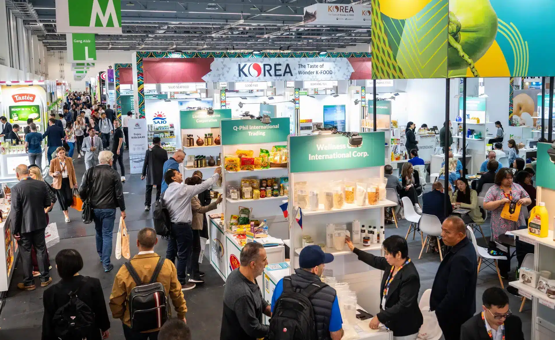 Eine geschäftige Messe oder Ausstellung voller Besucher, die zwischen Ständen wechseln und verschiedene internationale Lebensmittelprodukte präsentieren. Inmitten der Ausstellungen spiegelt ein markantes „Korea“-Banner die globale Repräsentation wider und erinnert an die lebhafte Atmosphäre der Anuga.