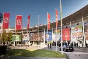 Außenansicht des belebten Eingangsbereichs des Kongresszentrums. Leuchtend rote Fahnen mit weißen Anuga-Logos schmücken das Bild. Besucher tummeln sich zwischen Bäumen und parkenden Autos. Unter dem klaren Himmel über ihnen werben Banner für verschiedene internationale Ausstellungen.