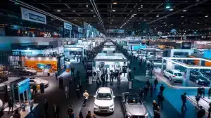 Eine geschäftige, gut beleuchtete Automobilausstellung, die an die Automechanika erinnert, bietet zahlreiche Besucher, die in einem großen Innenraum verschiedene Autoausstellungen erkunden. Dutzende Autos werden unter hellem Licht ausgestellt, und über den Messeständen sind Banner verschiedener Marken zu sehen.