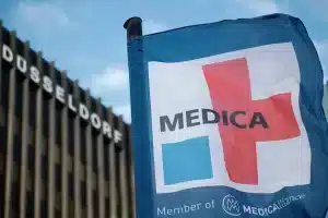 Eine Fahne mit der Aufschrift „MEDICA“ und einem Kreuz-Logo weht vor einem Gebäude mit der Aufschrift „DÜSSELDORF“ im klaren Himmel. Das Design weist auf die Zugehörigkeit als „Member of MEDICAlliance“ hin und unterstreicht die starke Verbindung zur Medica Düsseldorf.